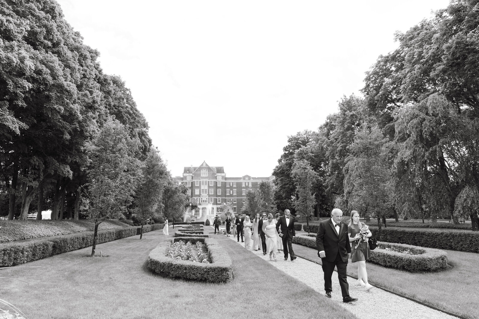 Black and white image of wedding party walking through European gardens toward estate, representing destination weddings by Clar Barron Photo.