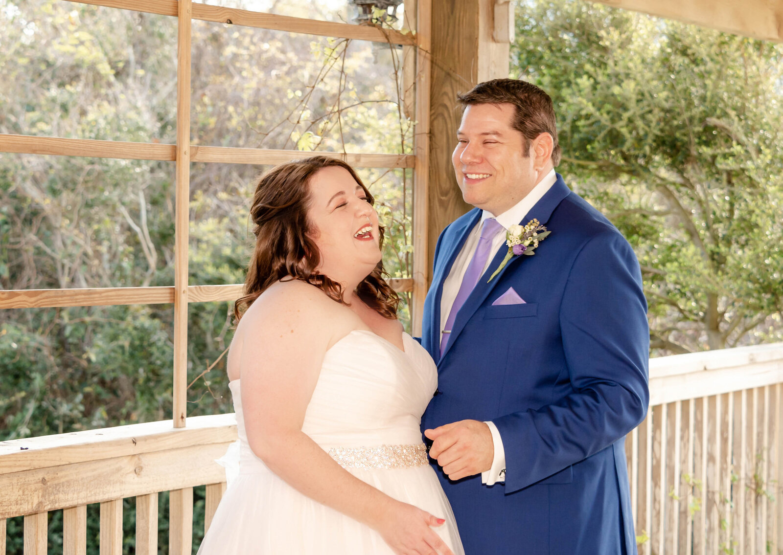 The bride and groom share a joyful moment together on a rustic porch with greenery in the background, captured during a Wrightsville Beach, NC wedding, highlighting authentic and candid wedding moments.