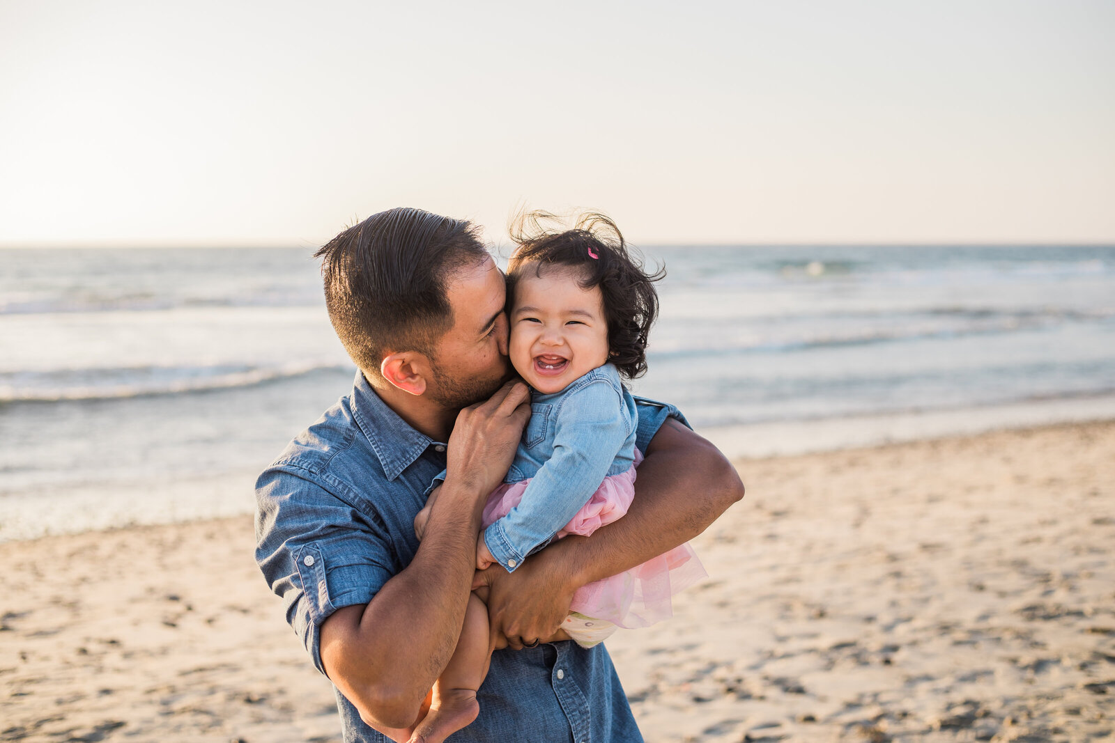 Family Photographer, a father kisses his giggling daughter at the beach