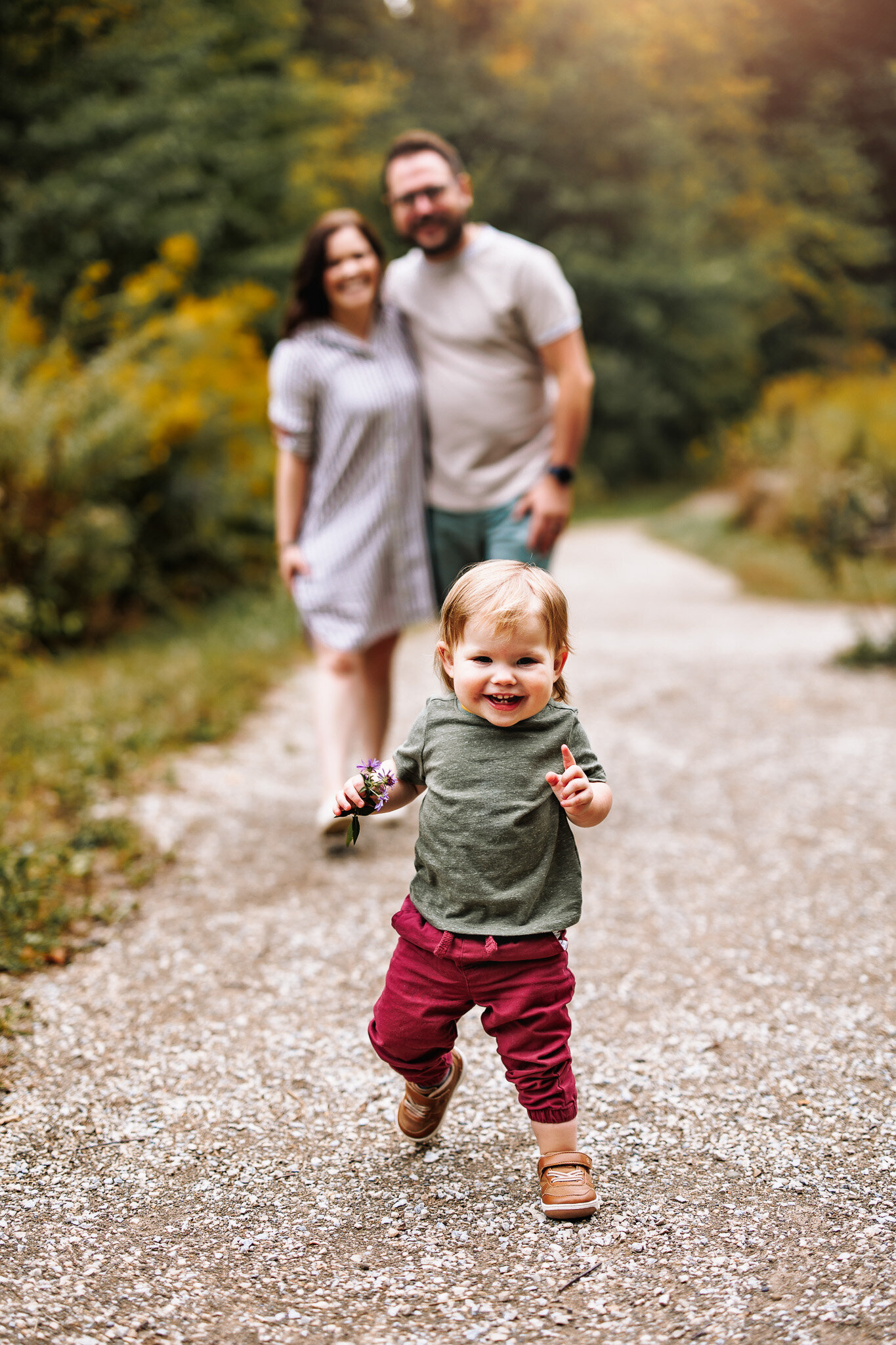 Along gravel path, A toddler boy runs towards the camera,  his mother and father holding each other close and smiling in the background