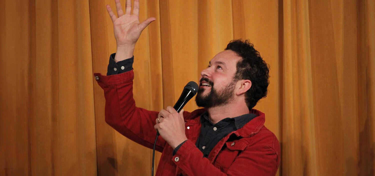 Comedian, Ahmed Bharoocha, holding a microphone and standing in front of an orange curtain background
