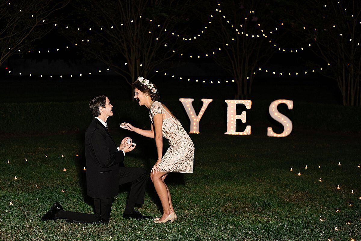 man on one knee wearing a black suit proposes to his fiancee wearing a short gold dress and flower crown in front of a light up YES sign