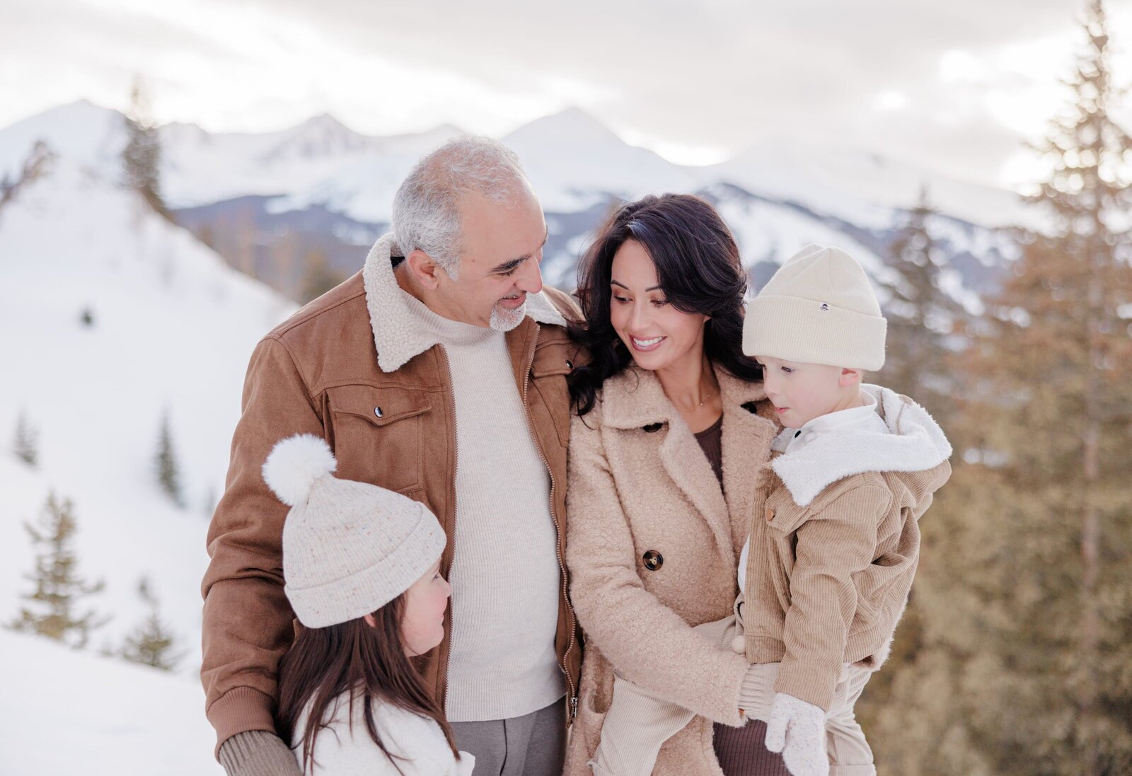 A family of four with two young children are huddled together on a winter afternoon. They are bundled in hats and gloves and are all smiling at one another and having a great time with a snowy winter scene behind them. 