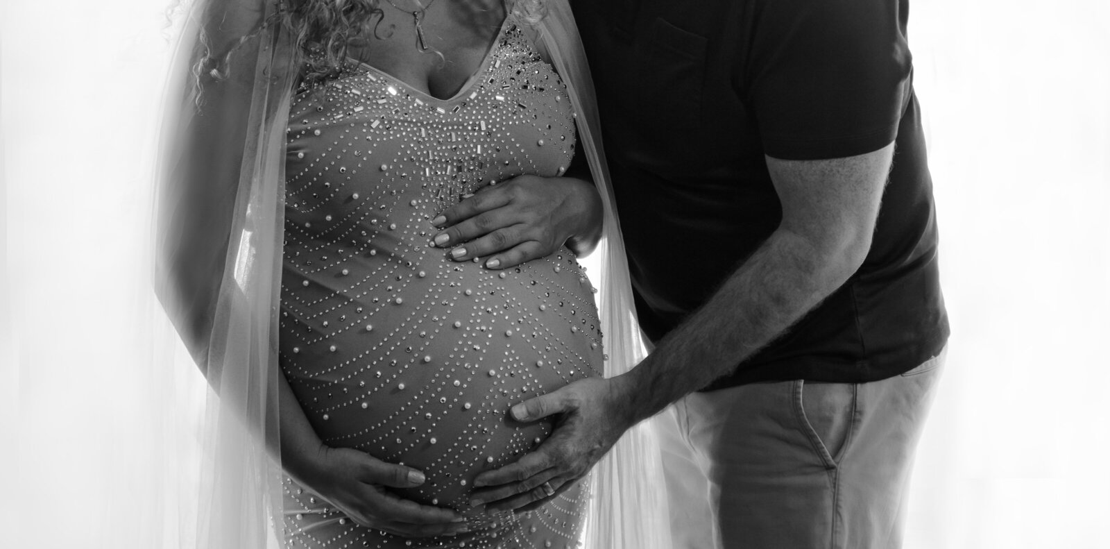 Glamorous maternity photo shoot captured by top Philadelphia and Main Line maternity and newborn photographer. Close up image of mom's bump. Mom is wearing a glamorous, fitted, beaded maternity gown. She has one hand at the top and bottom of her bump. Dad is standing with his chest facing mom and his forearm underneath her bump.
