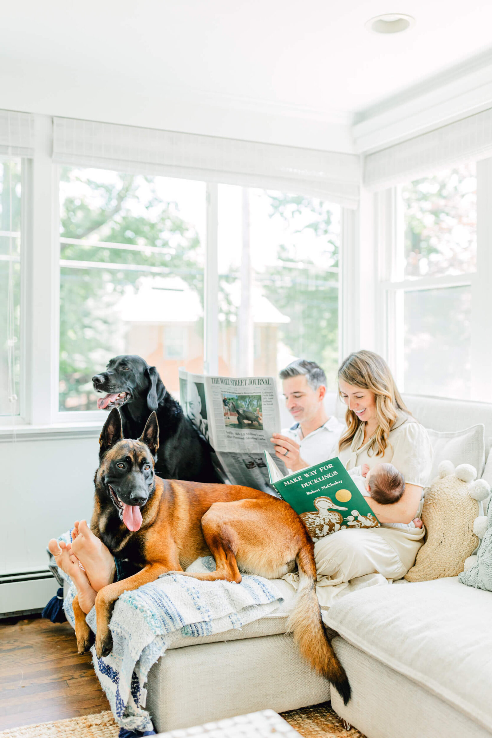 Mom and dad read next to each other on a couch with mom holding their newborn and two dogs sitting at their feet