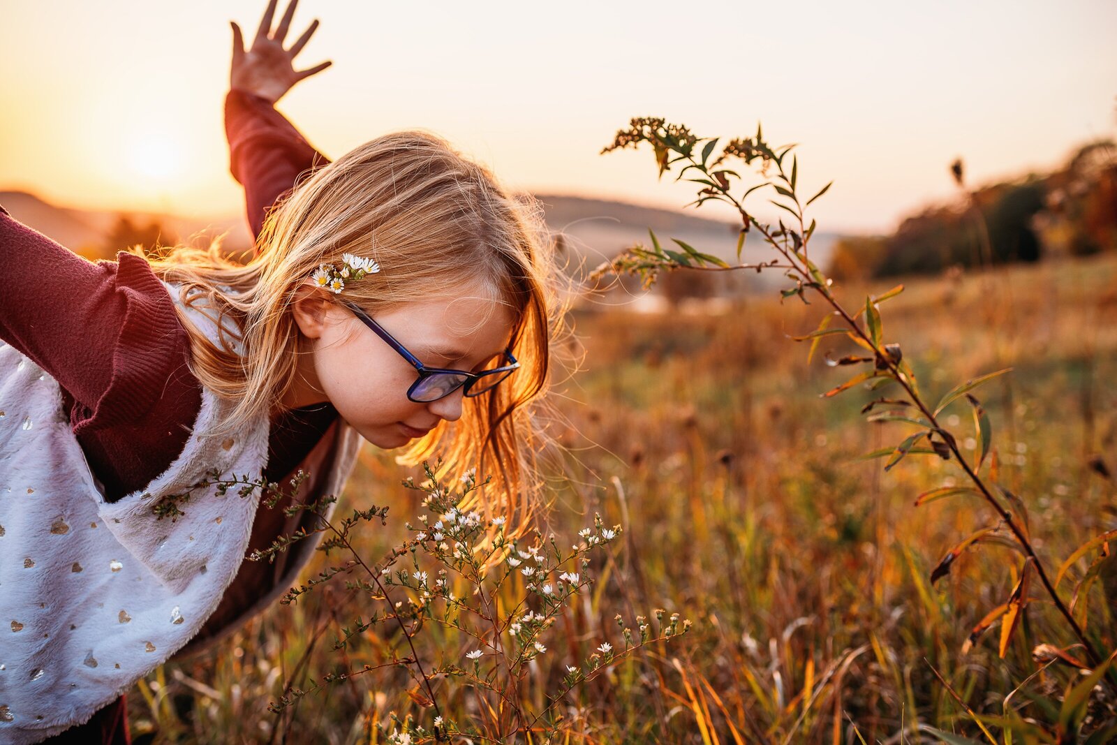Child with long hair and glasses leaning forward to smell flower, arms outstretched behind.