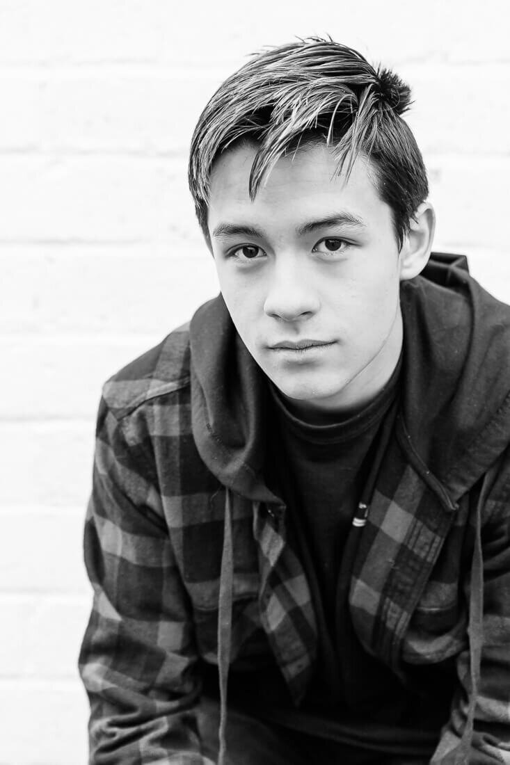 Salt Lake City senior black and white photography of a high school senior boy wearing a plaid jacket sitting in front of a white brick wall in Lehi in the Spring