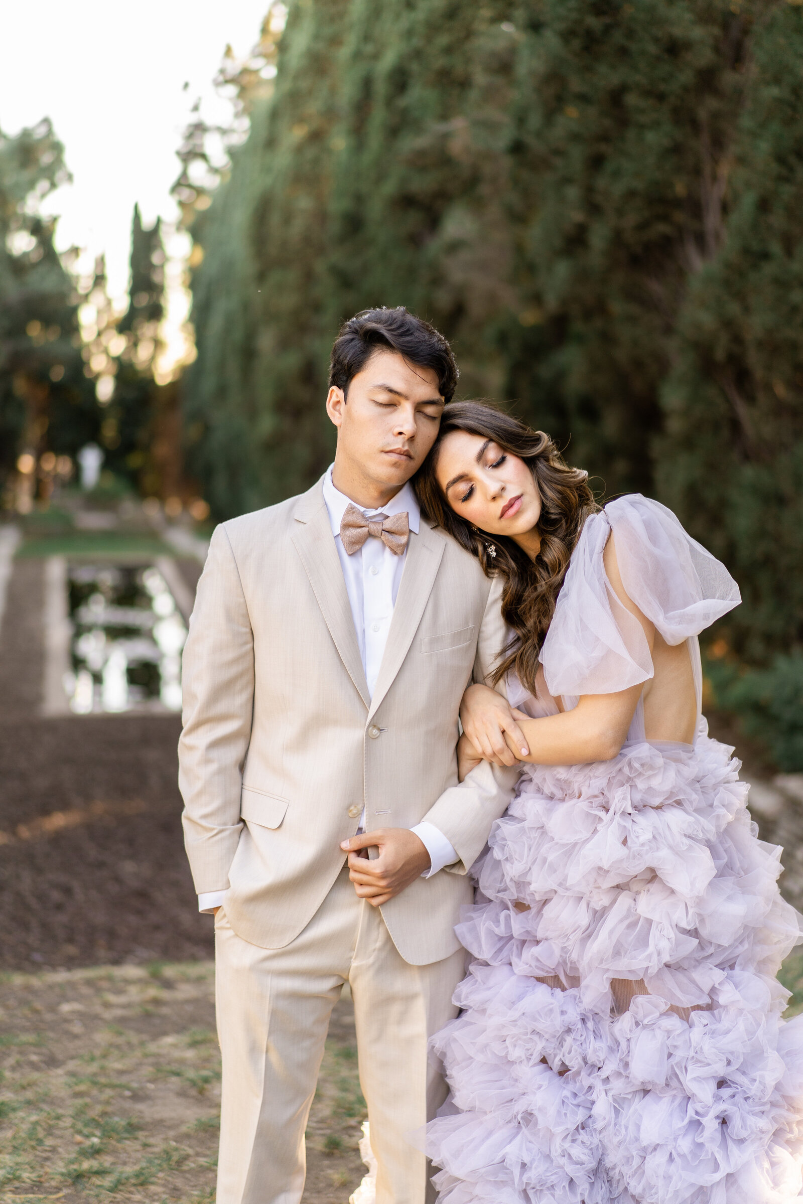 Portrait of bride and groom in a lavender gown and cream suit standing near a water structure and large trees.