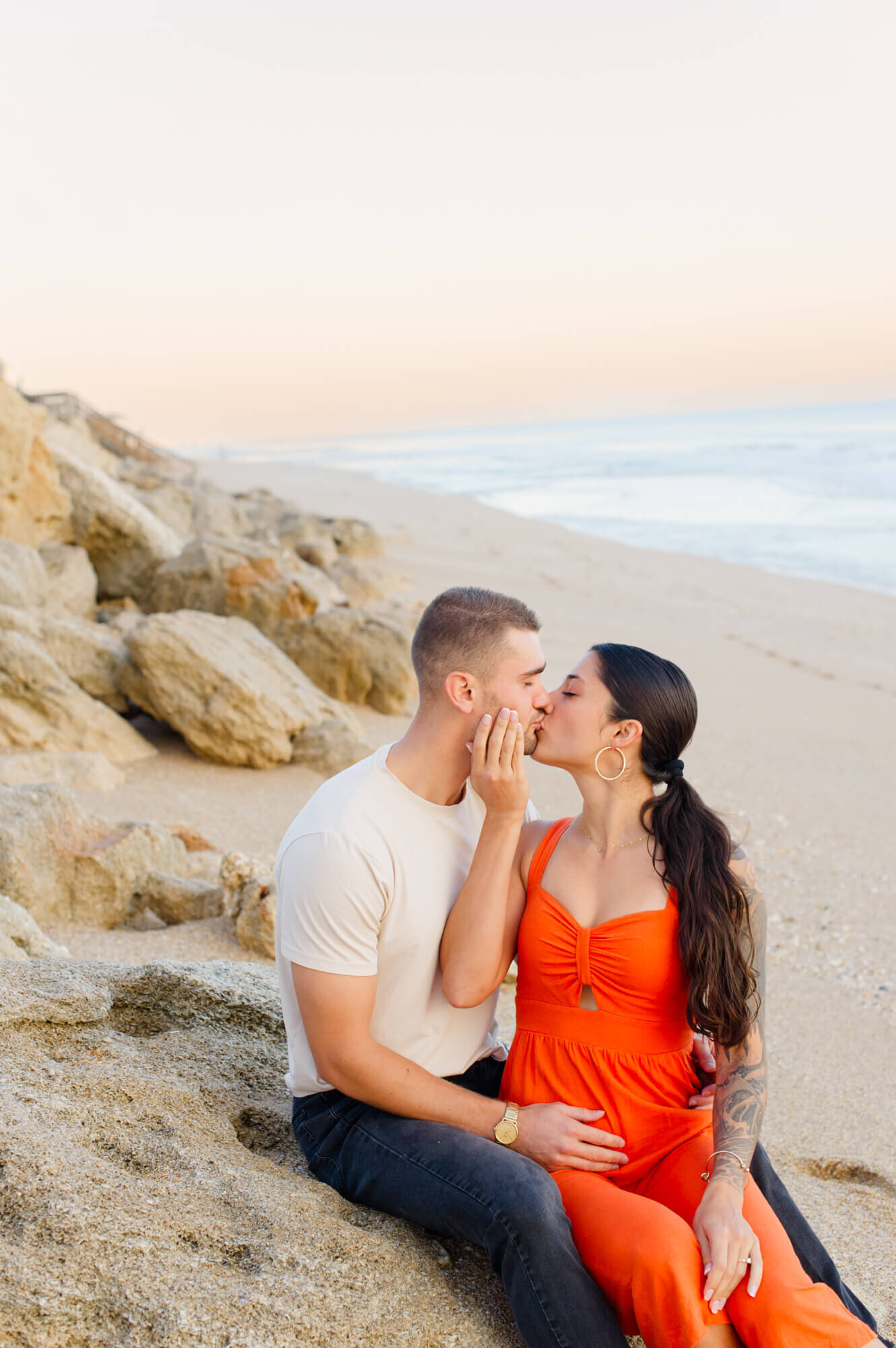 Newly engaged couple sits on rocks kissing during their  beach engagement photo session