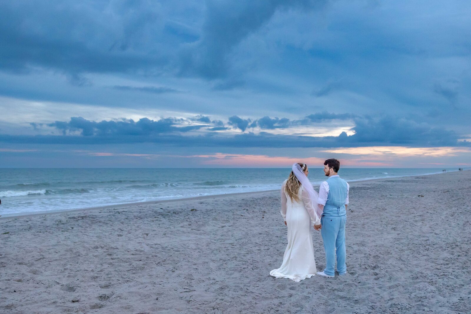 The bride and groom stand together on the beach at sunset, holding hands and gazing at the horizon, captured during a Topsail Beach, NC wedding, blending romance with natural coastal beauty.