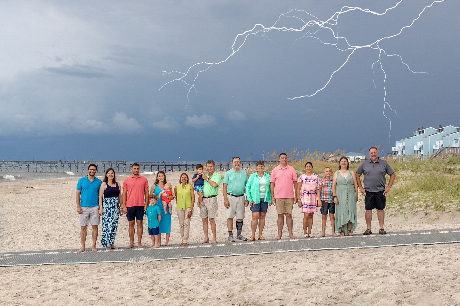 A large family stands on a beach boardwalk with dramatic lightning illuminating the sky. Dressed in colorful outfits, they capture a striking and memorable moment. Perfect for North Carolina family photography, highlighting dynamic beach scenes.