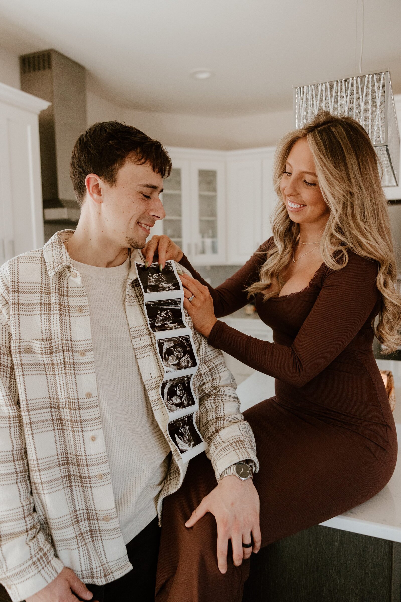 A joyful couple shares a sweet moment in their modern kitchen while holding ultrasound photos. The mom-to-be, dressed in a fitted brown dress, playfully pins the sonogram strip to her partner’s plaid shirt as they smile at each other, celebrating their pregnancy announcement.