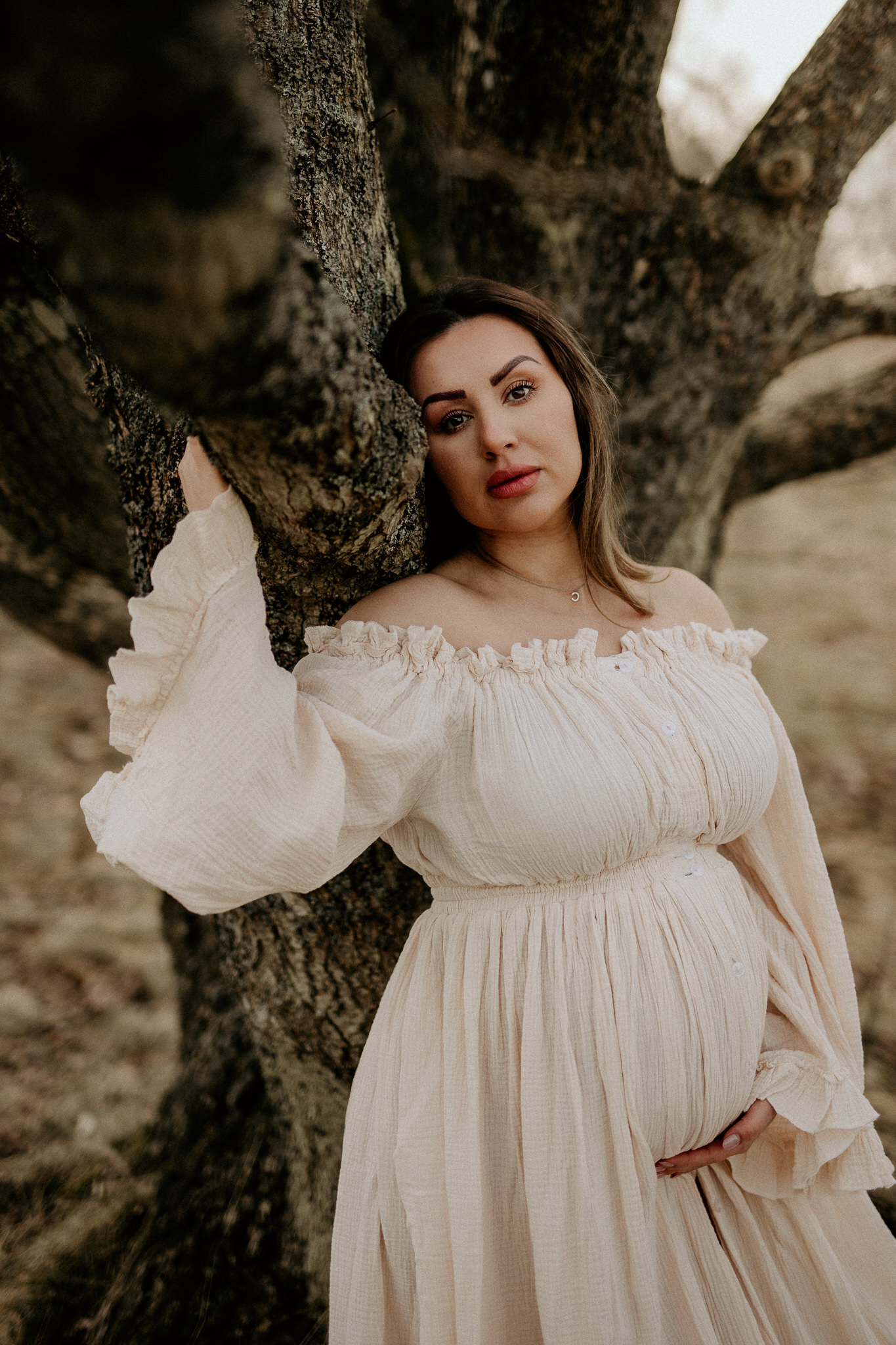 en gravid kvinna står i en bohemisk gravidklänning och lutar sig mot ett träd