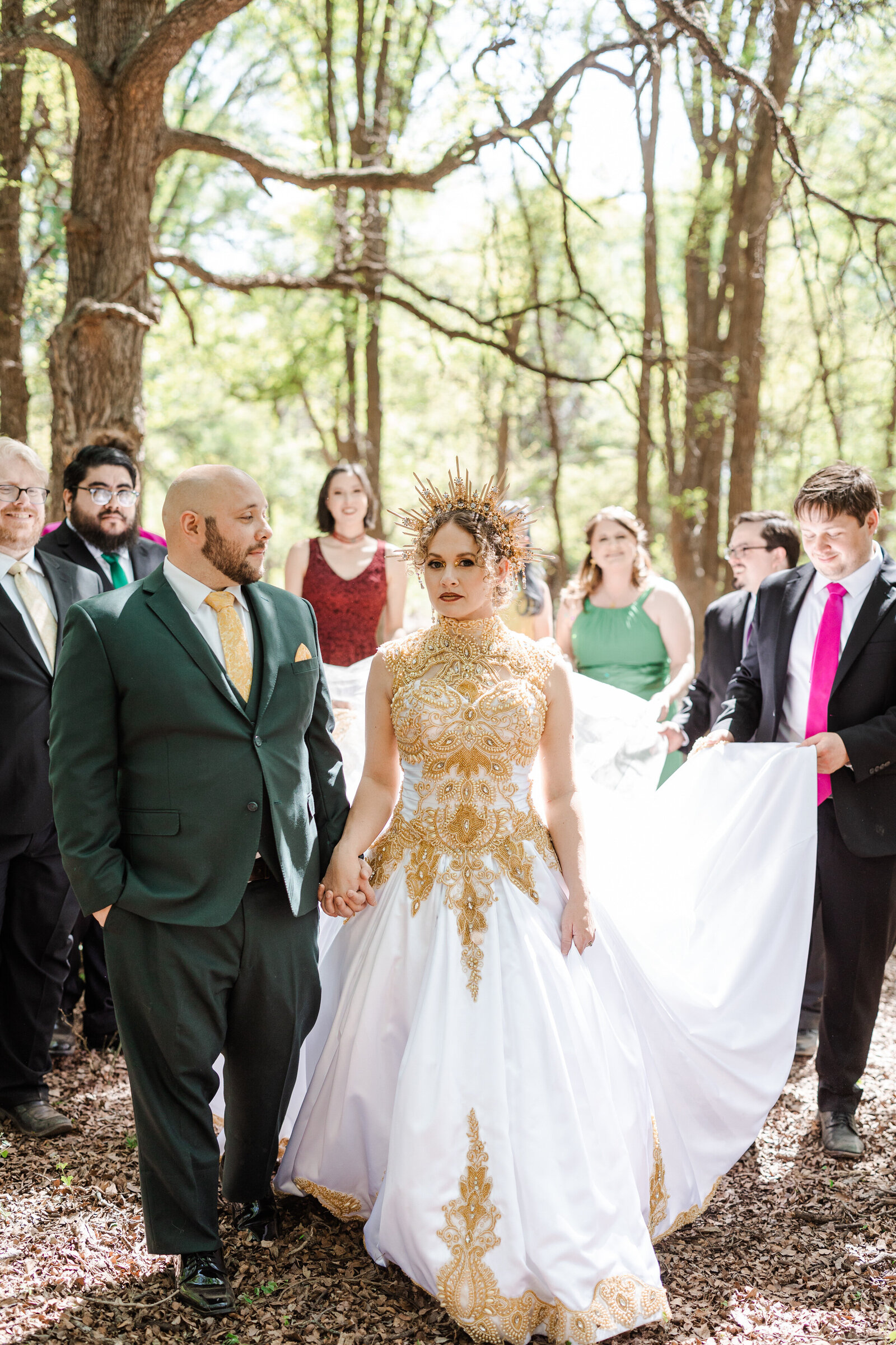 A bride in a gold crown and gold accented dress holds her groom's hand while her wedding party fans out her long dress train behind her. The groom is wearing a green suit and is looking at the bride. The wedding party all follow joyfully behind them in a wooded setting.