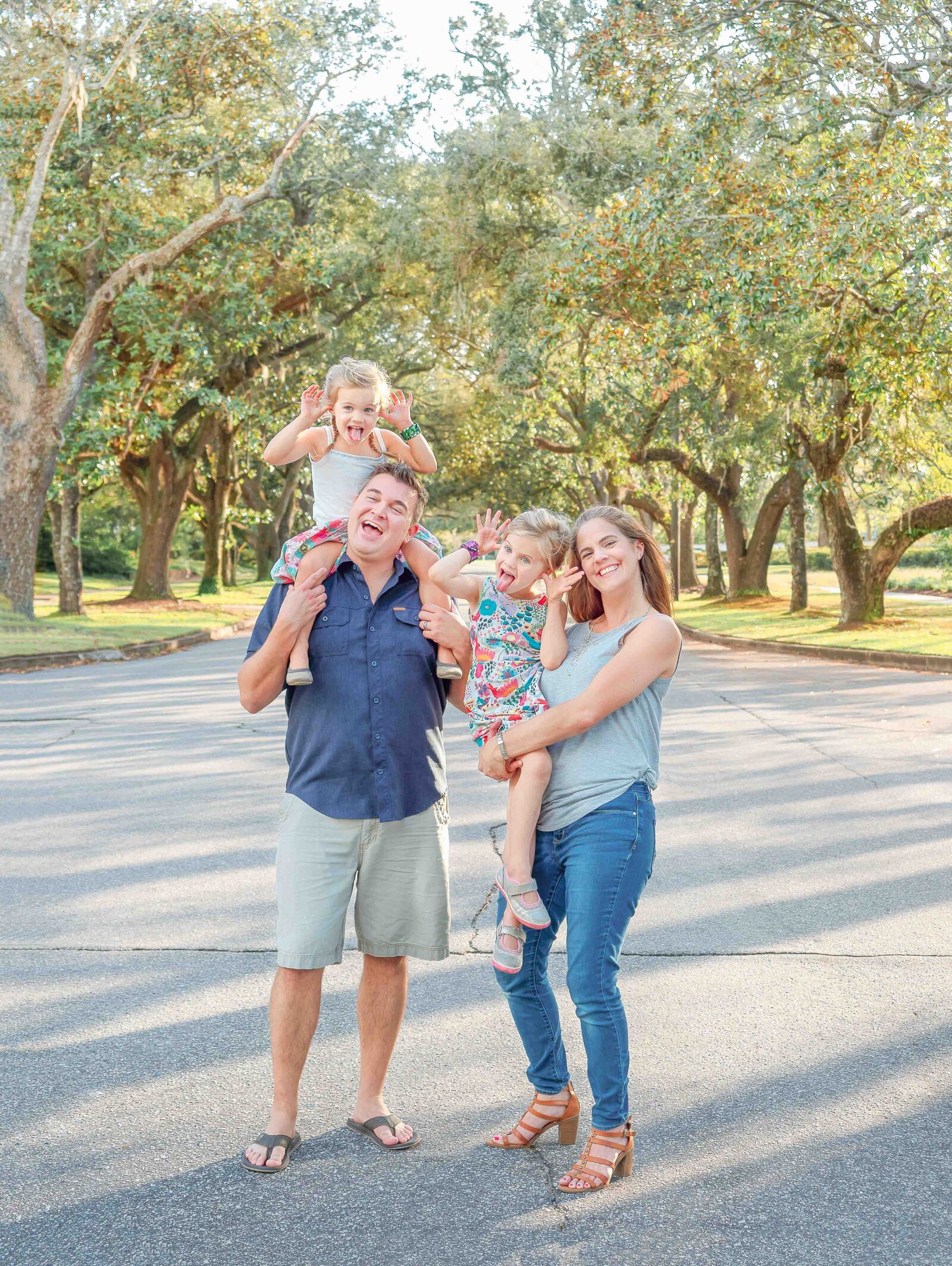 A playful family poses in a lush, tree-lined park near Wilmington, NC, with two young children on their parents' shoulders. The dappled sunlight and vibrant greenery create a lively atmosphere. This image exemplifies destination family photography in NC, ideal for capturing candid, joyful moments.