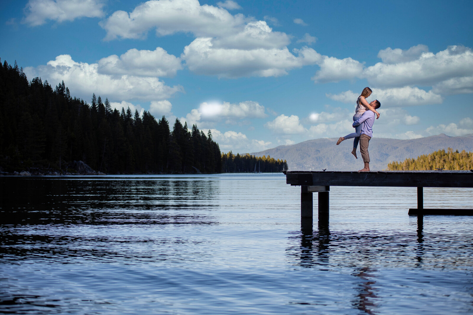 Engaged couple on a dock in emerald bay lake tahoe with the lake and trees in the background. The male is holding up his fiance in his arms as she kicks one leg out and looks into his eyes, photo by wedding photographer sacramento ca, philippe studio pro.