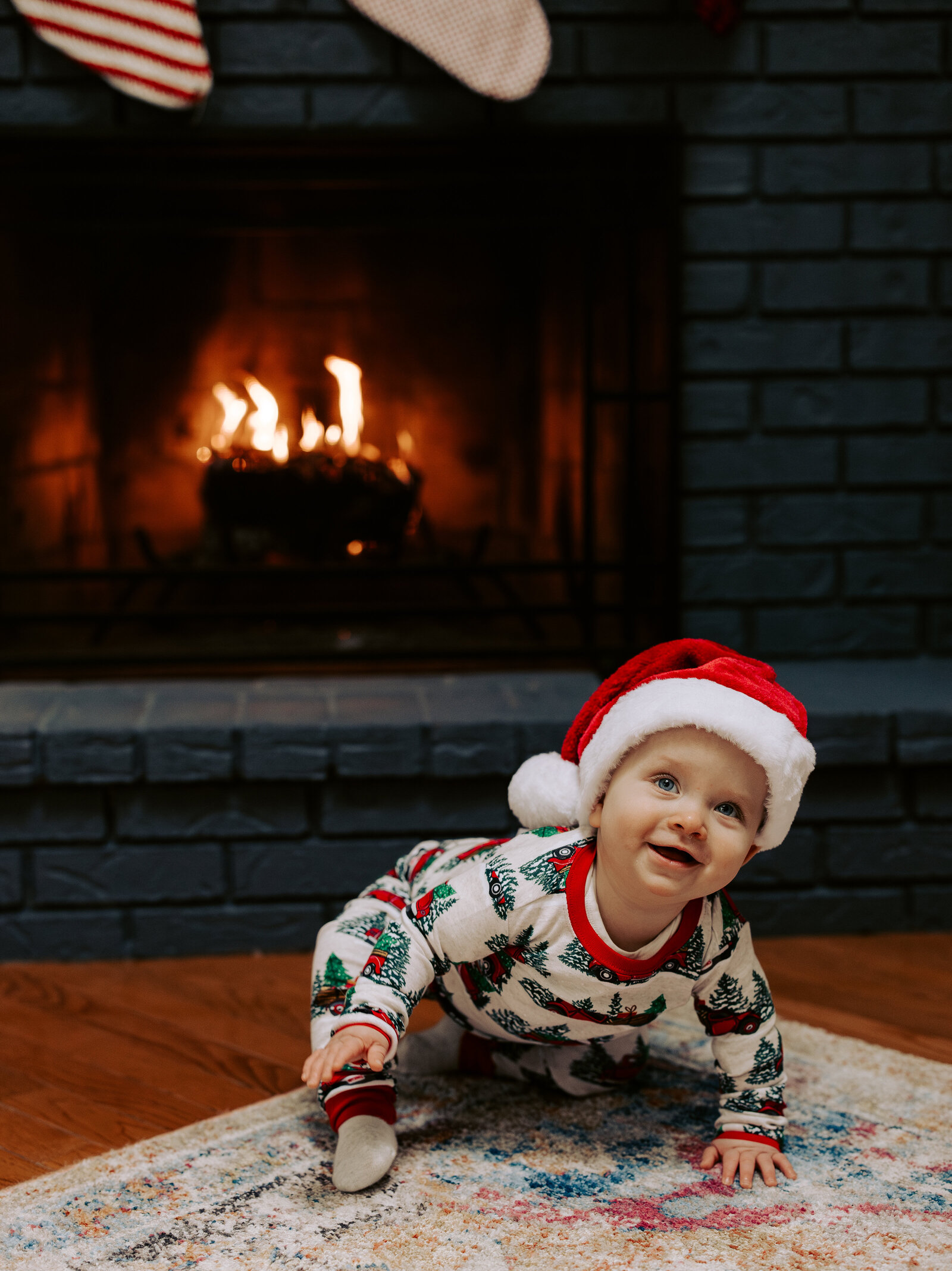 Baby in santa hat smiles in front of fireplace during her winter family photo session in Londonderry, New Hampshire