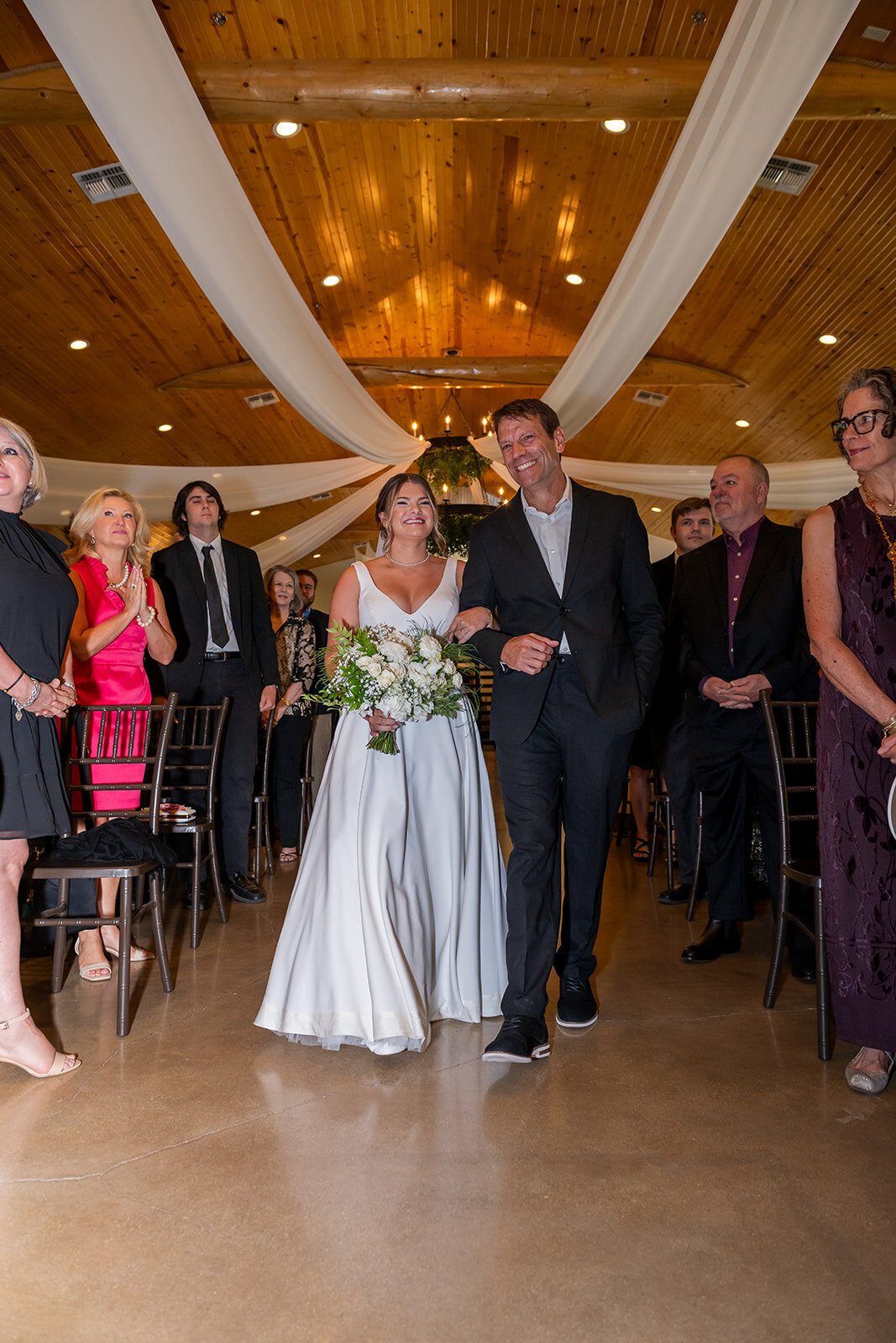 A beautiful moment as the bride walks down the aisle with her father, surrounded by loved ones in a warmly lit ceremony venue. Captured by CMH Studios in San Antonio.