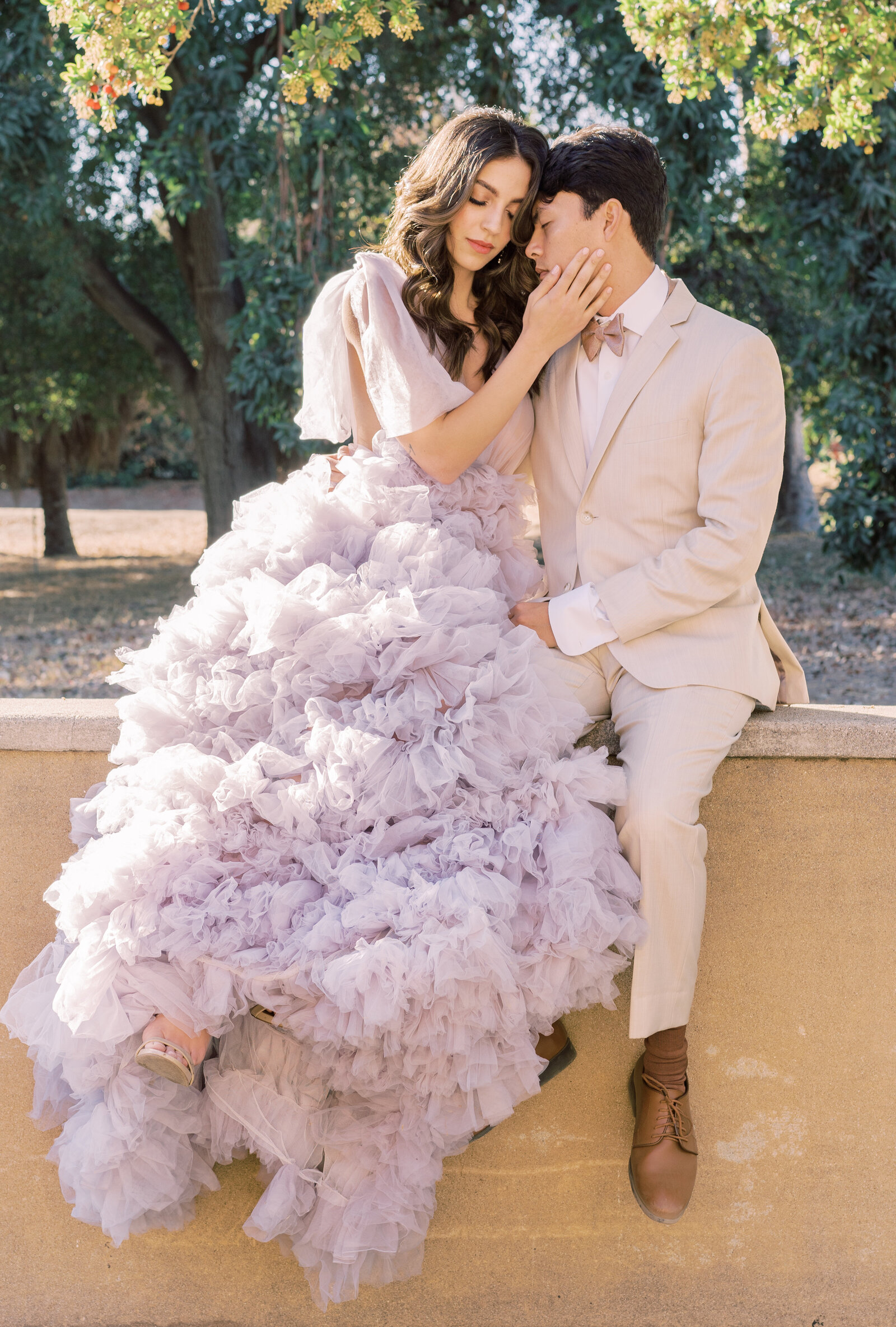 Portrait of bride and groom in a lavender gown and cream suit sitting on a peach-colored wall with trees in the back.