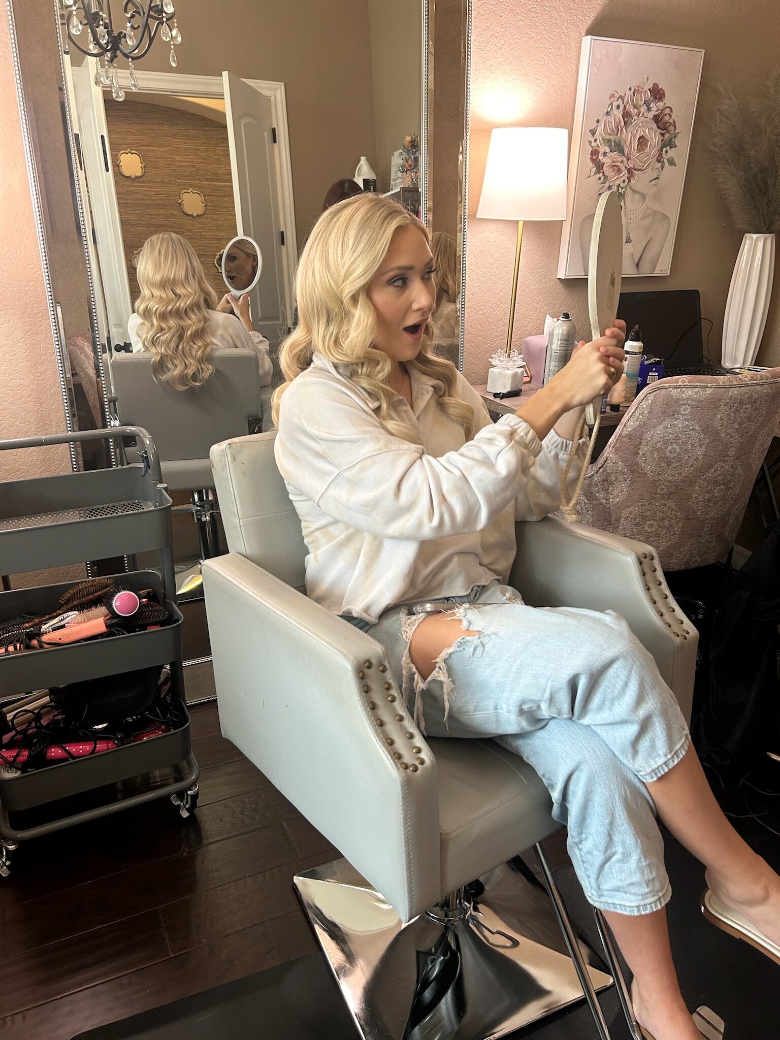 A woman sits in a salon chair, holding a mirror and looking surprised at her reflection with styled hair. It's all part of the bridal beauty experience Texas brides rave about, where wedding beauty consultations reveal just what to expect from a wedding makeup artist on your big day.