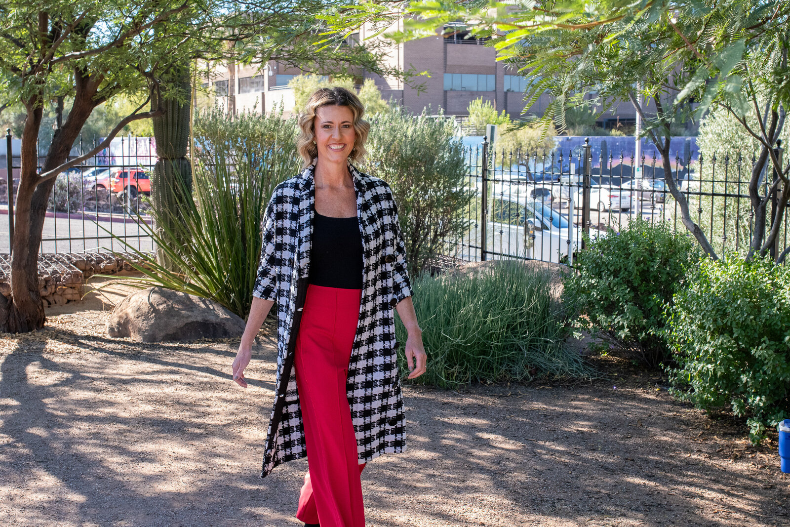 Woman walking confidently through a sunny garden area wearing red pants and a patterned long coat, photographed by Vyrl Photo, showcasing brand photography Tucson.