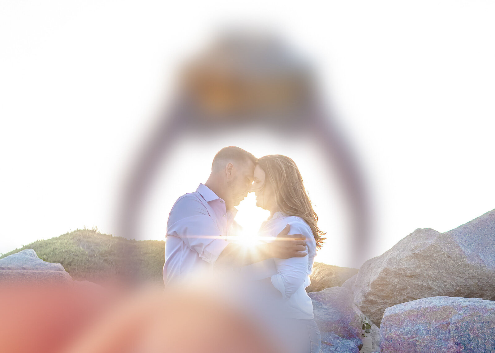 A romantic couple stands closely together on a rocky hillside, with sunlight shining through their embrace. The shot is artistically framed through a wedding ring, symbolizing love and commitment. This creative composition highlights the breathtaking style of Bella Lumiere Photography, a North Carolina wedding company. 