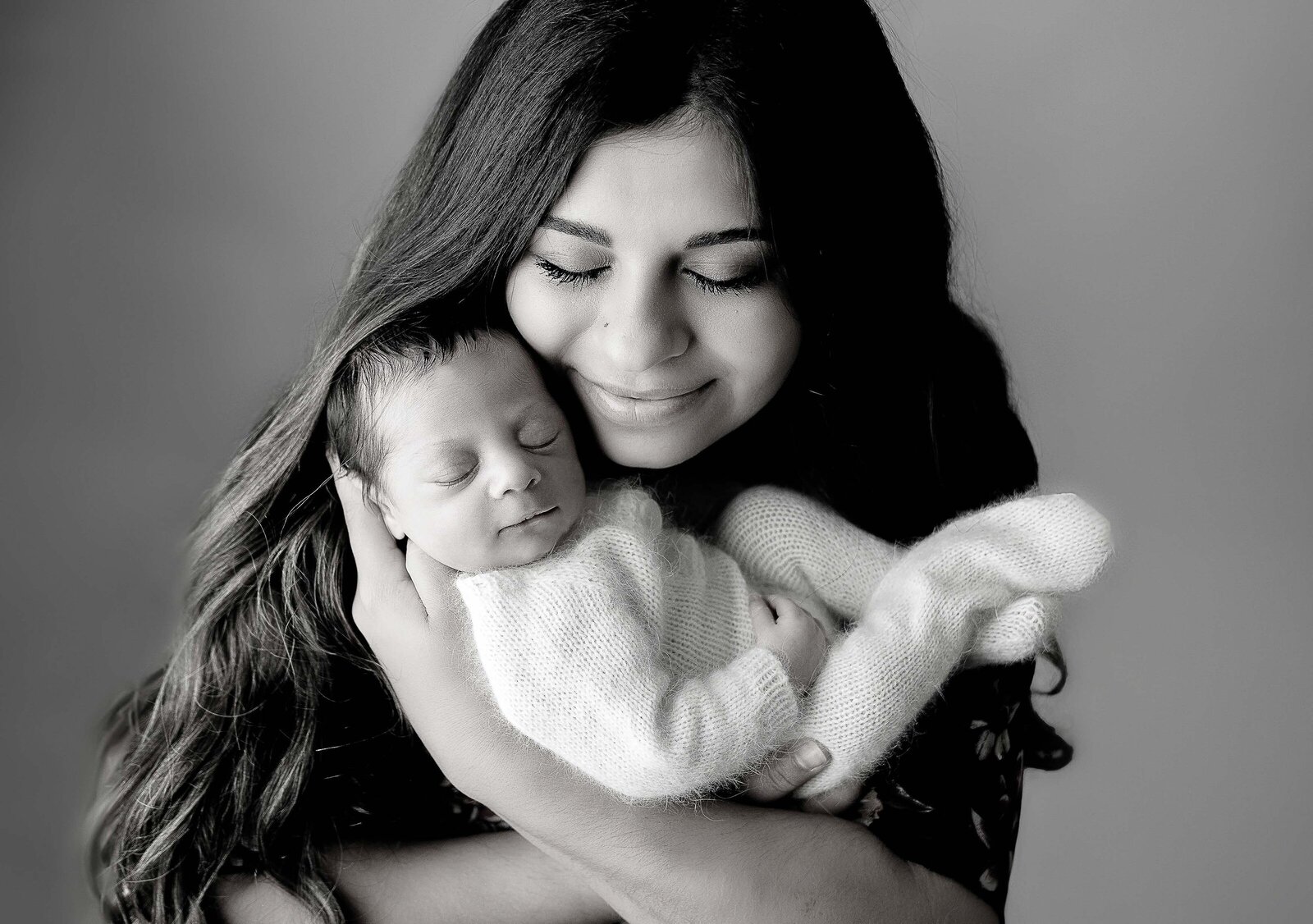 Newborn studio photo shoot with top Philadelphia and Main Line newborn photographer. Mom is cradling baby in her arms against her upper chest. Her eyes are closed and resting her cheek against baby's temple. Baby's eyes are close and sleeping peacefully.