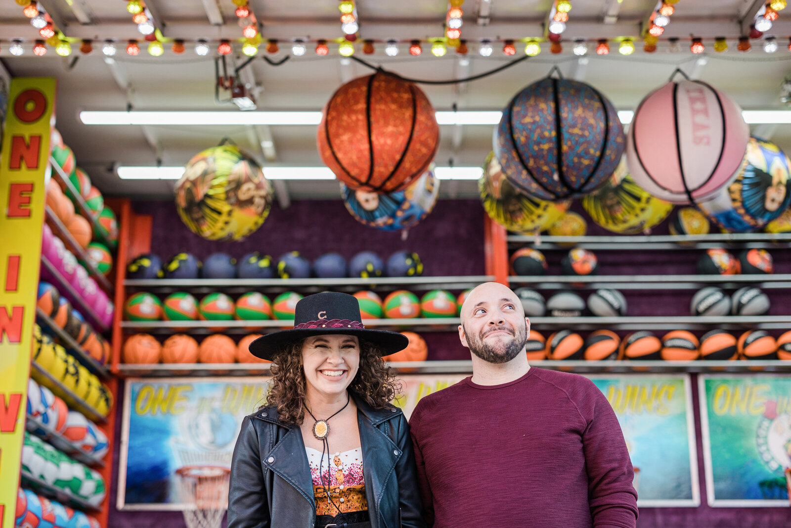 A couple playfully posing in front of a midway game during their carnival engagement session in Fort Worth, Texas. The woman on the left is wearing a black, brimmed hat, colorful dress, and a black leather jacket while smiling and looking directly at the camera. The man on the right is wearing a maroon sweater and is smiling coyly while looking off to the right with only his eyes.