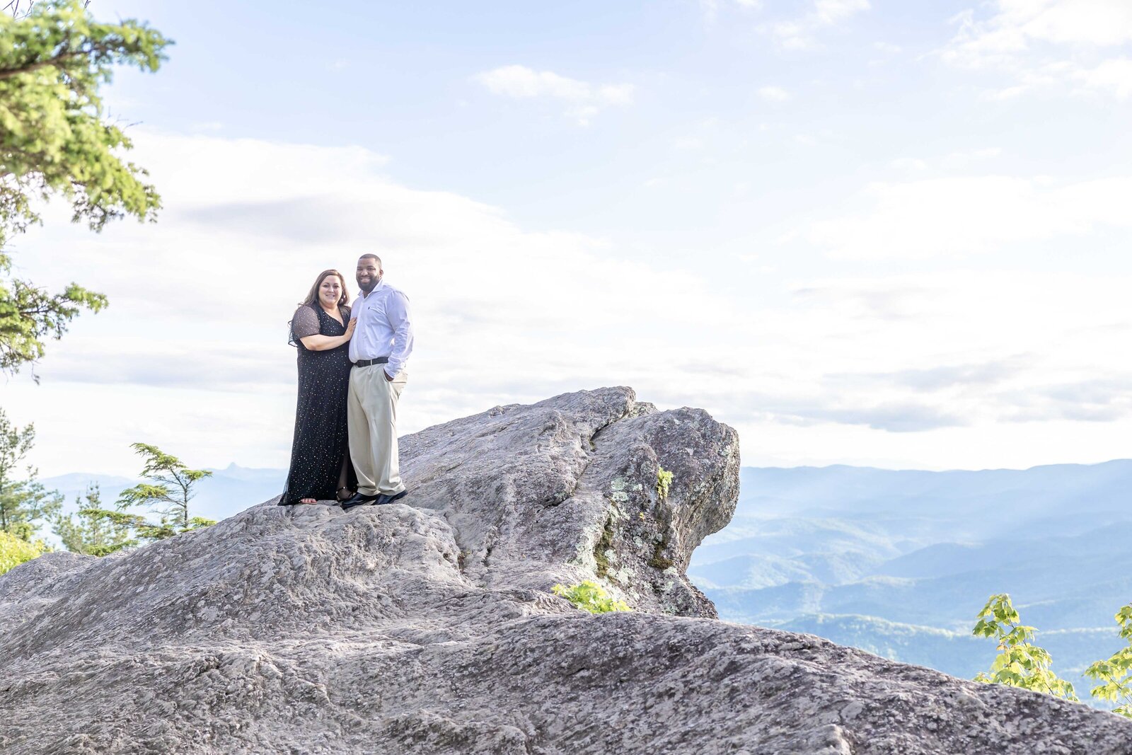 A couple stands on the dramatic Blowing Rock mountain top with breathtaking views of rolling hills and a bright sky in the background. The woman wears a long black dress, and the man is dressed in a light shirt and trousers. This scenic moment beautifully showcases the adventurous spirit of North Carolina engagement photography.  