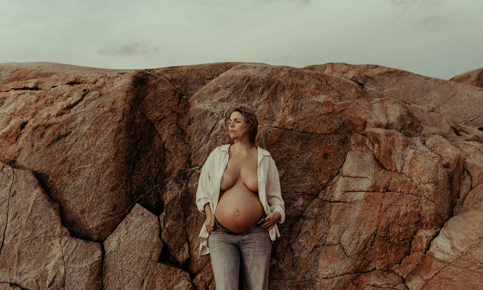 en gravid mamma i vit skjorta och jeans lutar sig mot klippor