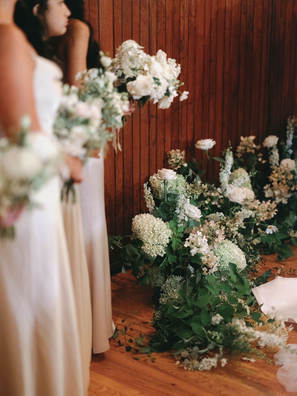 Rain plan Inside ceremony in Evergreen Museum carriage house with a lush white floral ground arch and bridesmaids in the foreground holding white bouquets.