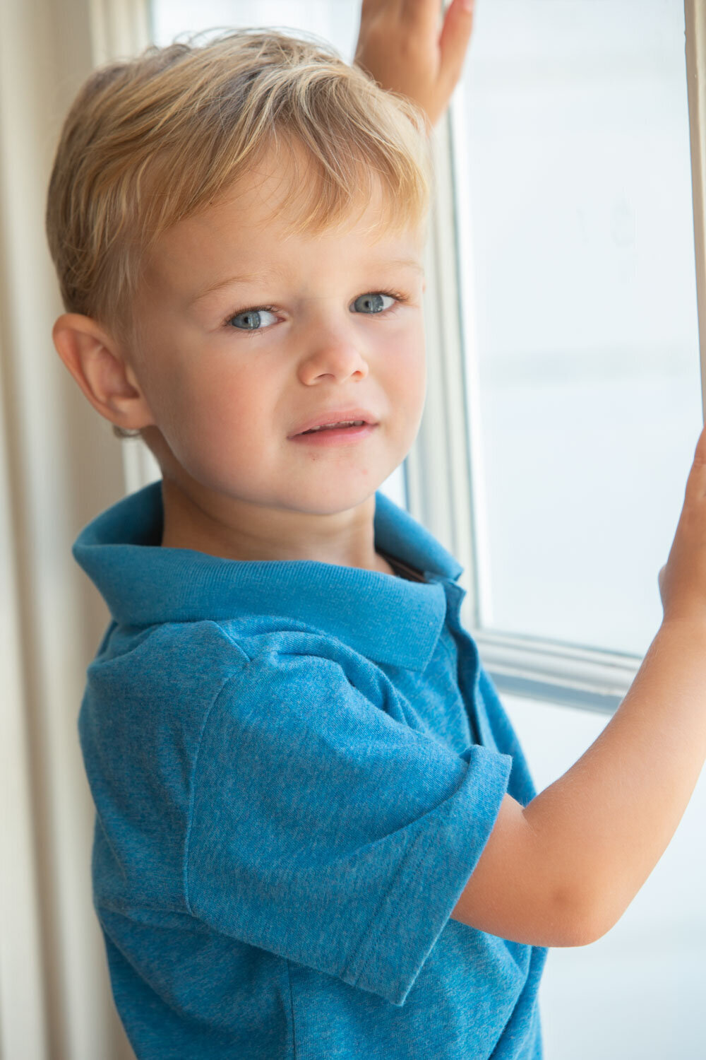 A young boy in a blue shirt is looking out a window