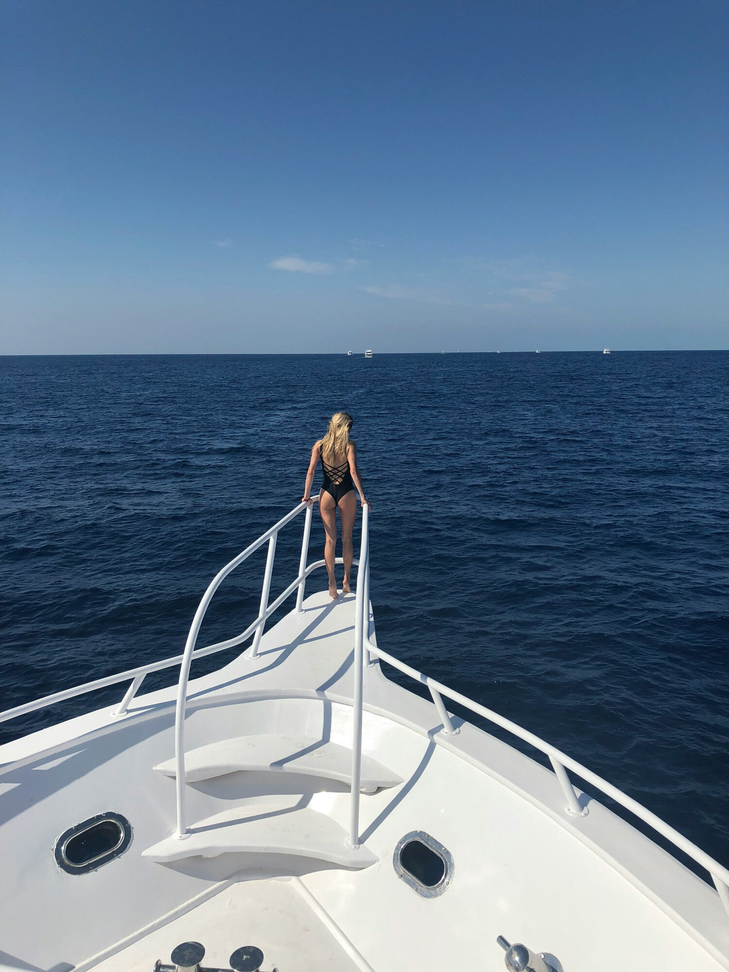 A person in a black swimsuit standing at the bow of a white boat, facing the open sea under a clear blue sky
