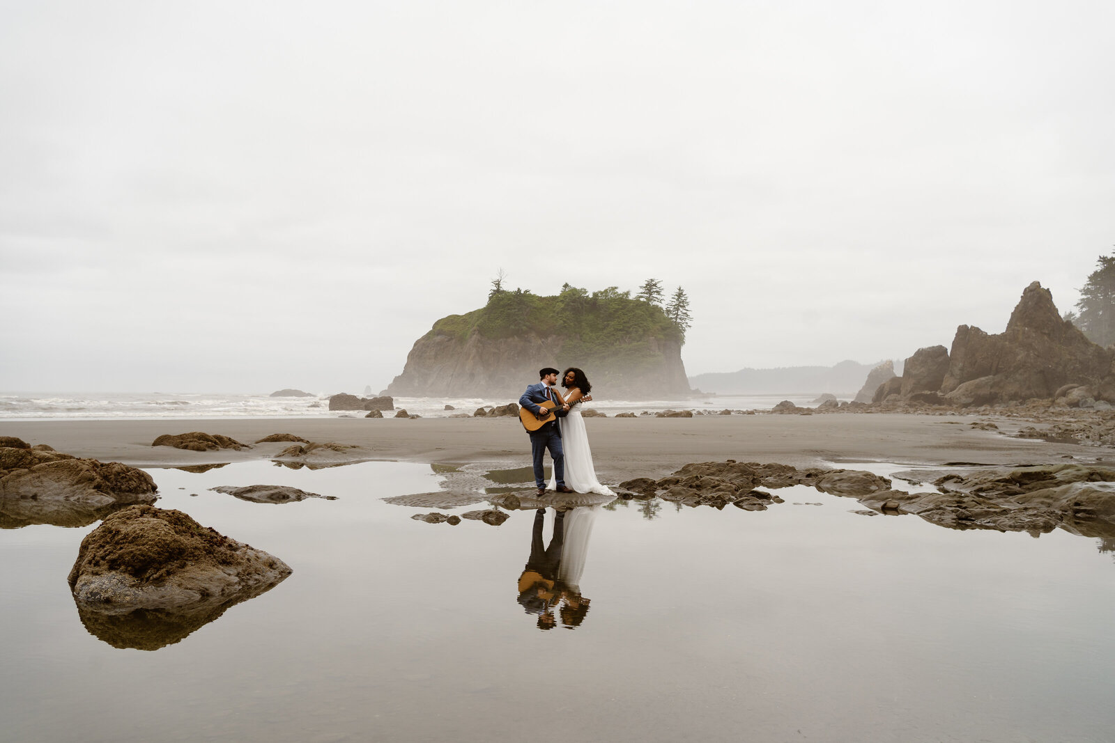 a couple is reflected in the water of ruby beach, the groom plays guitar while a bride embraces him from behind during their washington elopement