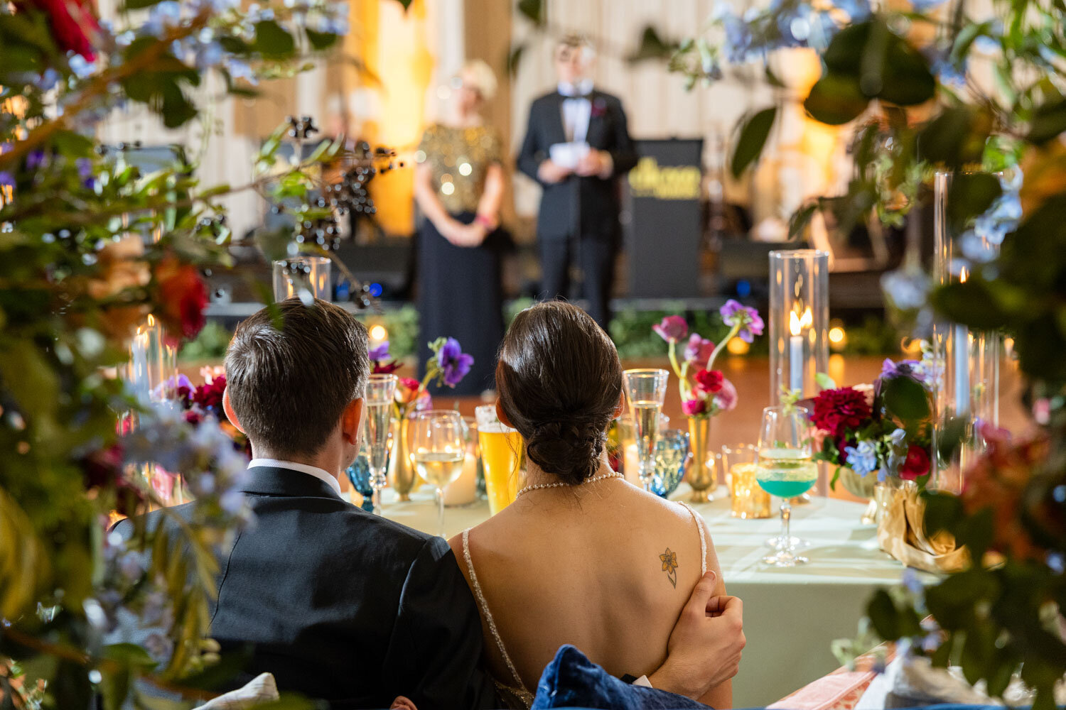 A bride and groom sit at a table with flowers and candles