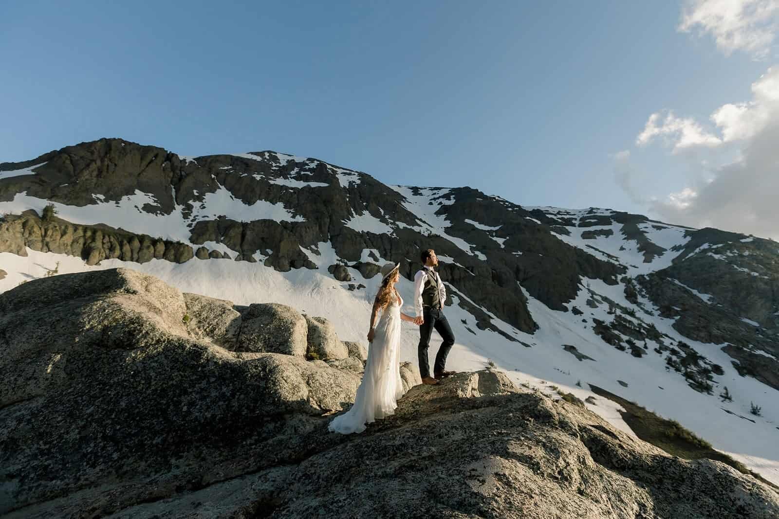 A bride and groom stand in front of snow covered mountains at high elevation on the Sonora Pass on their wedding day. 