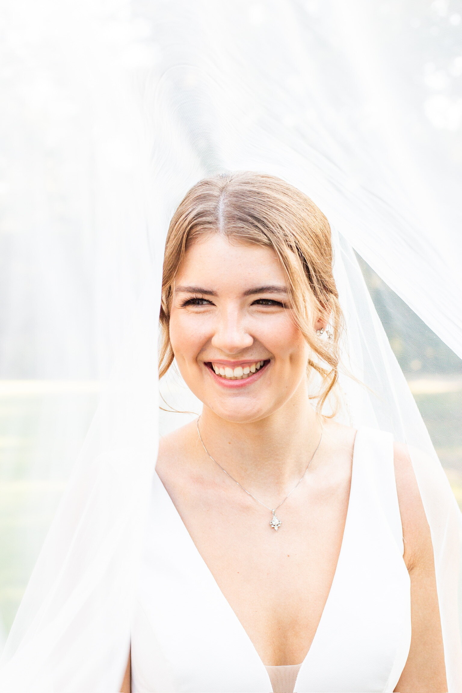 Bride smiling under a sheer veil, wearing a white dress with a V-neck, standing outdoors, embodying the bridal beauty experience Texas offers through expert wedding makeup artistry.