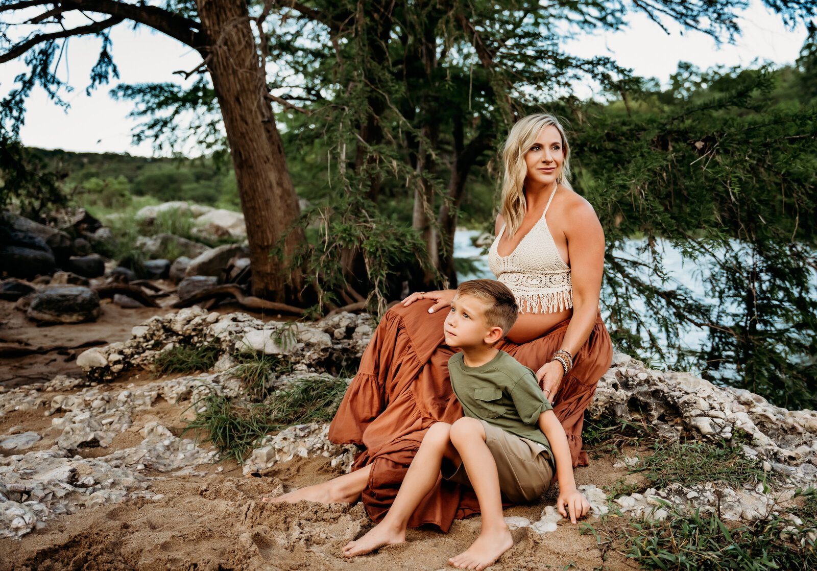 Maternity Photographer, a mother-to-be sits on a rock in the forest with her young son