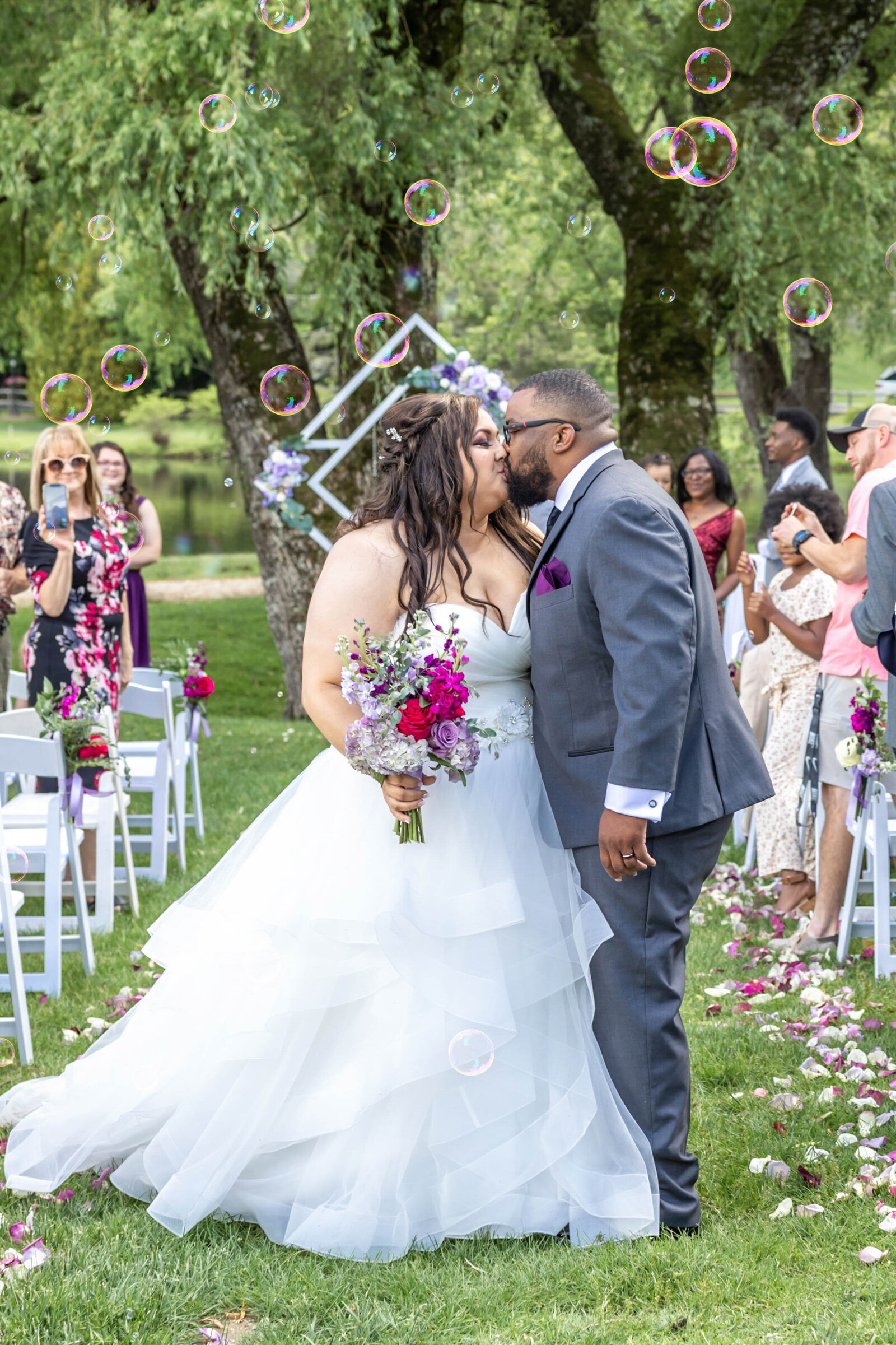 The bride and groom kiss under floating bubbles during their outdoor wedding ceremony, surrounded by lush greenery and colorful florals, captured during a Boone, NC wedding, blending romance and natural beauty.