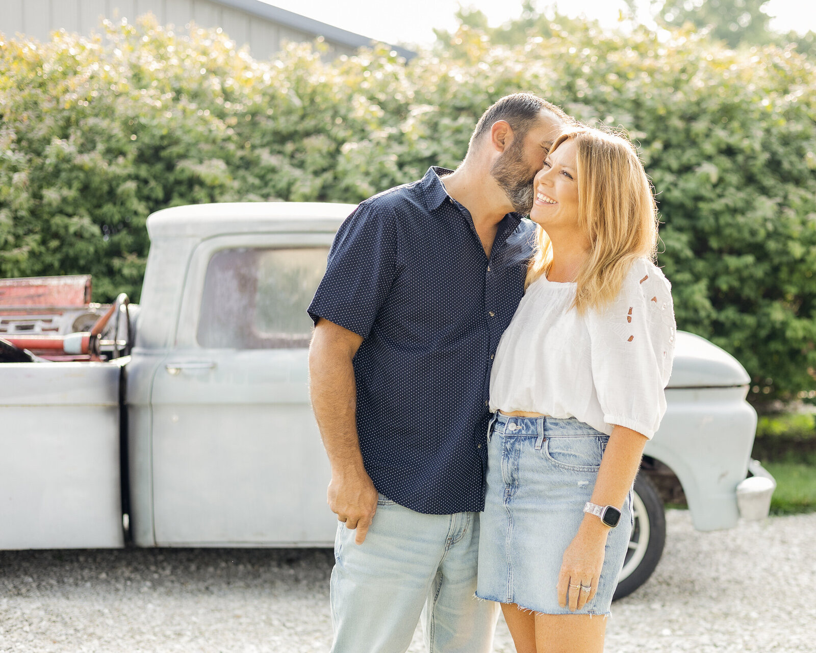 Husband and wife standing in front of a classic truck, husband is leaning into his wife's ear, has dark hair and beard, wearing a dark blue button up shirt, and blue jeans.  Wife has blond hair, wearing a white mid sleeve shirt with a blue jean skirt, smiling.