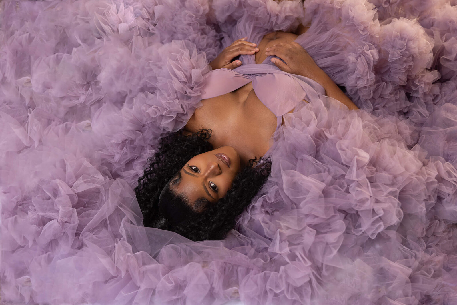 An expectant mother lies in a sea of soft lavender tulle, her serene expression capturing the beauty of maternity.