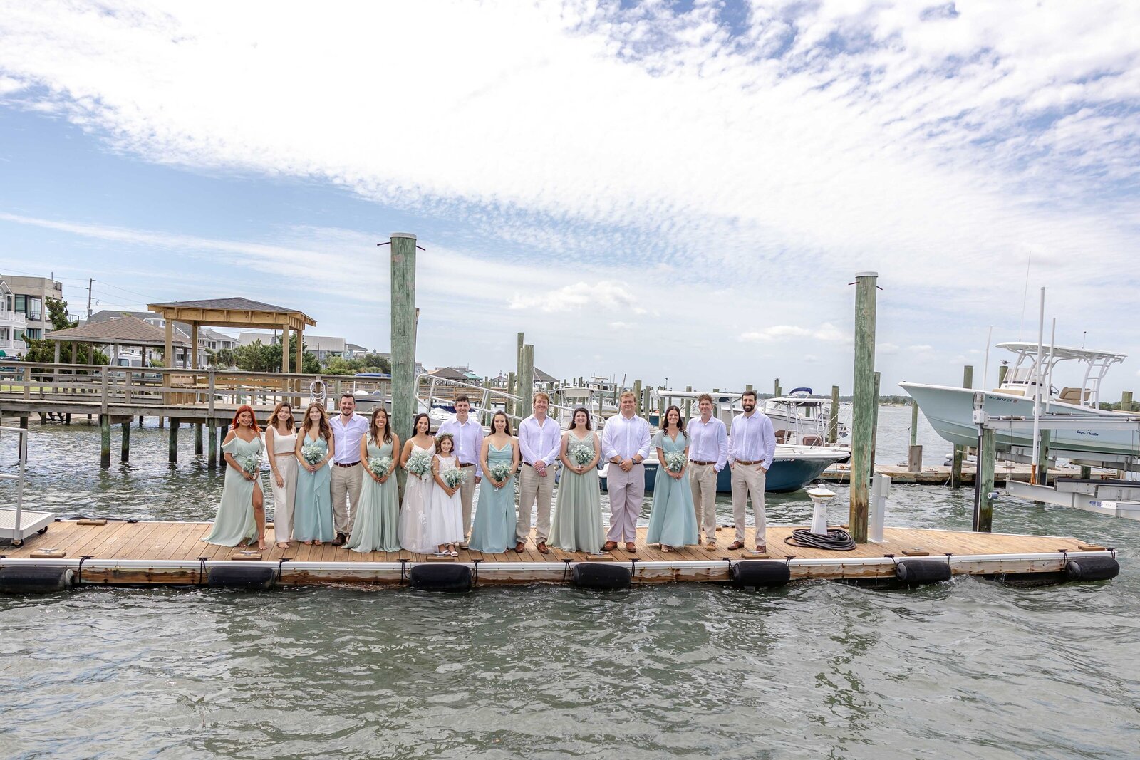 A wedding party in sage green and white attire poses on a wooden dock with boats and the waterfront in the background, captured at a Wrightsville Beach, NC wedding, showcasing coastal elegance and group celebration.