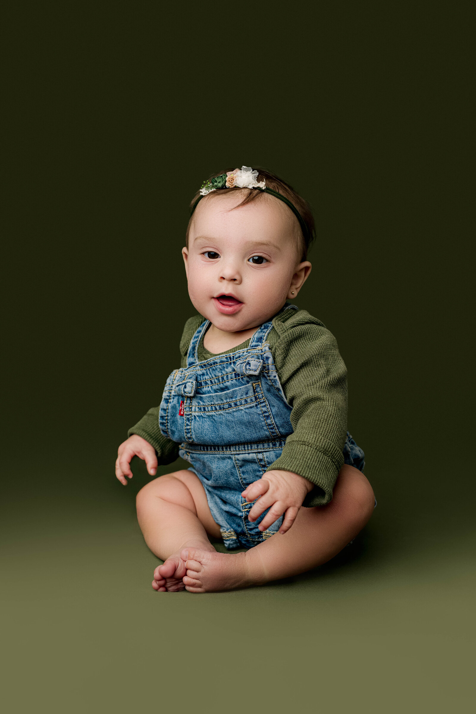 Baby girl in denim overalls and a green top, seated confidently in studio lighting during her milestone session in Charlotte.