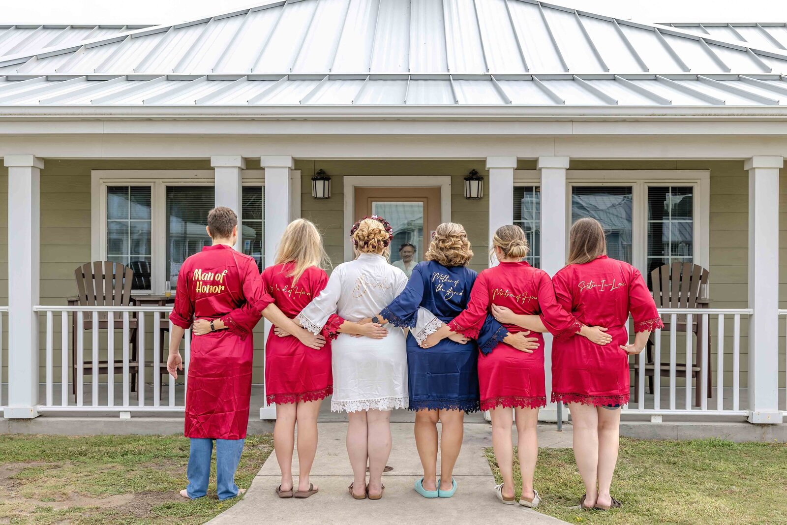 The bride and bridesmaids, dressed in personalized red and blue robes, pose with their arms around each other on a porch, captured during a Fort Fisher, NC wedding, highlighting joyful and relaxed pre-ceremony moments.