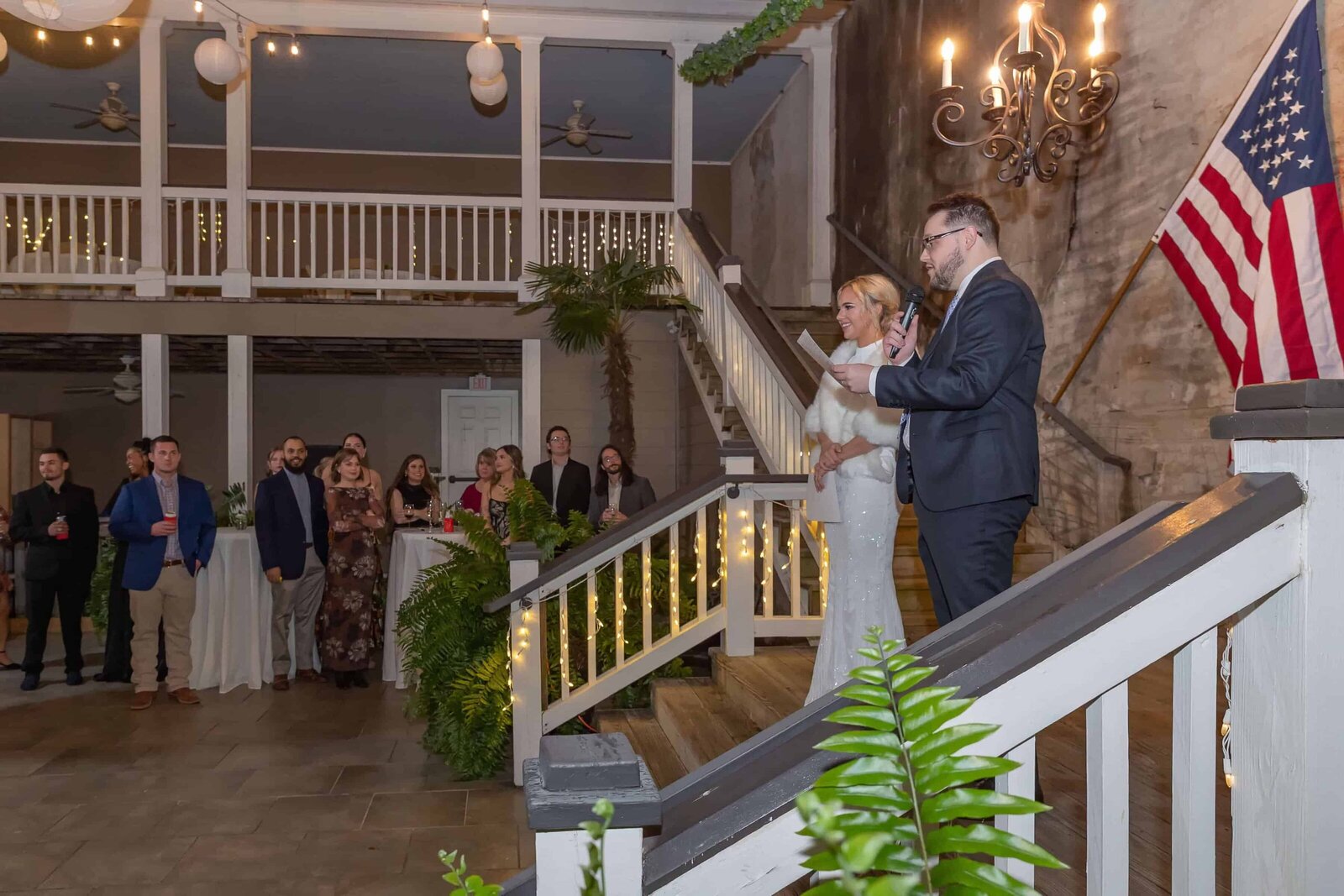A bride and groom stand on a staircase giving a speech at their wedding reception. Guests below listen attentively in a decorated venue with string lights, plants, and an American flag.