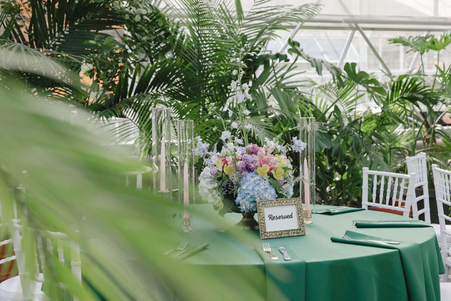 Table setup with green tablecloth and floral centerpiece in a lush, greenhouse-like setting