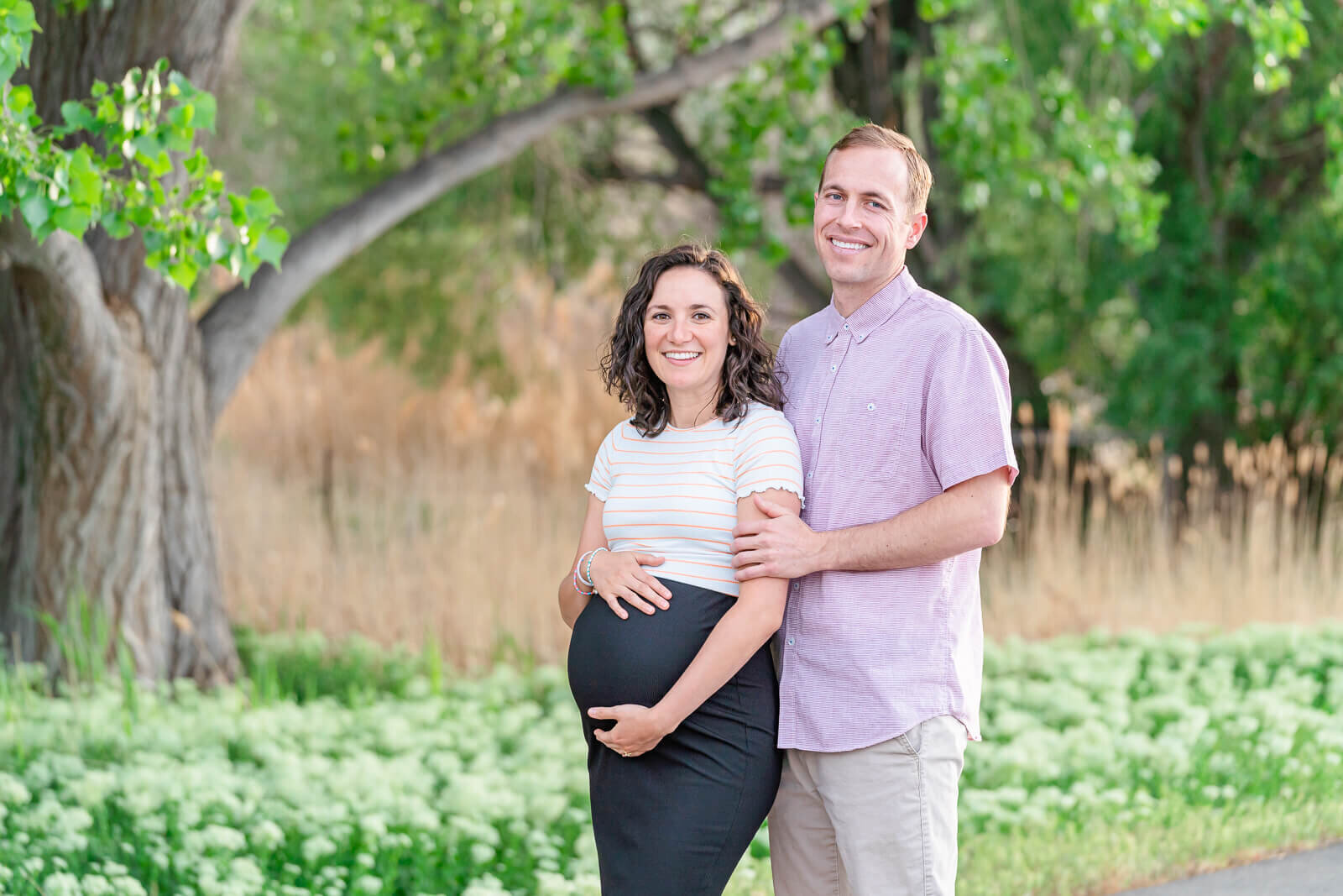 Utah maternity photography of a pregnant woman wearing a white striped shortsleeved top and a black skirt holds her belly with her husband wearing a pink shortsleeved button down shirt and khaki pants standing behind her