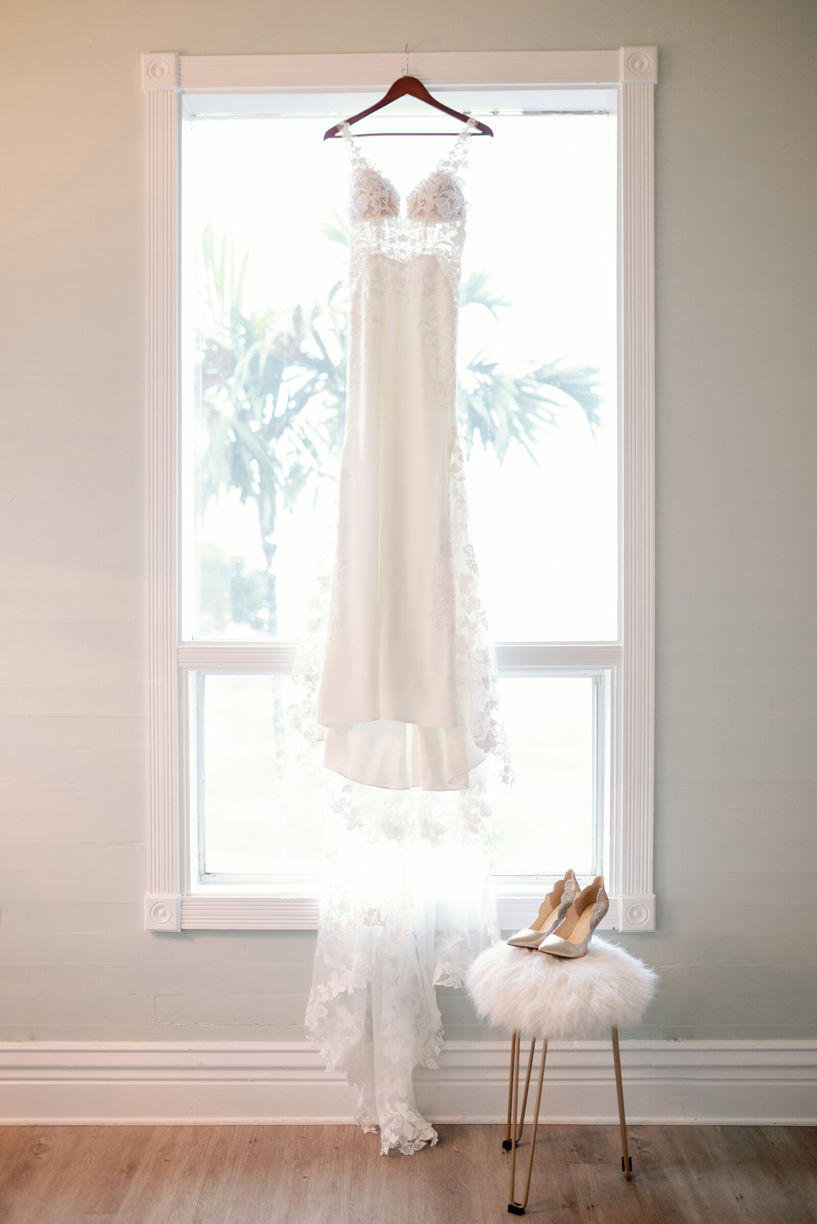 wedding dress hanging in a bright winder and wedding day shoes on a fluffy while and golden stool in a historic home in South Florida