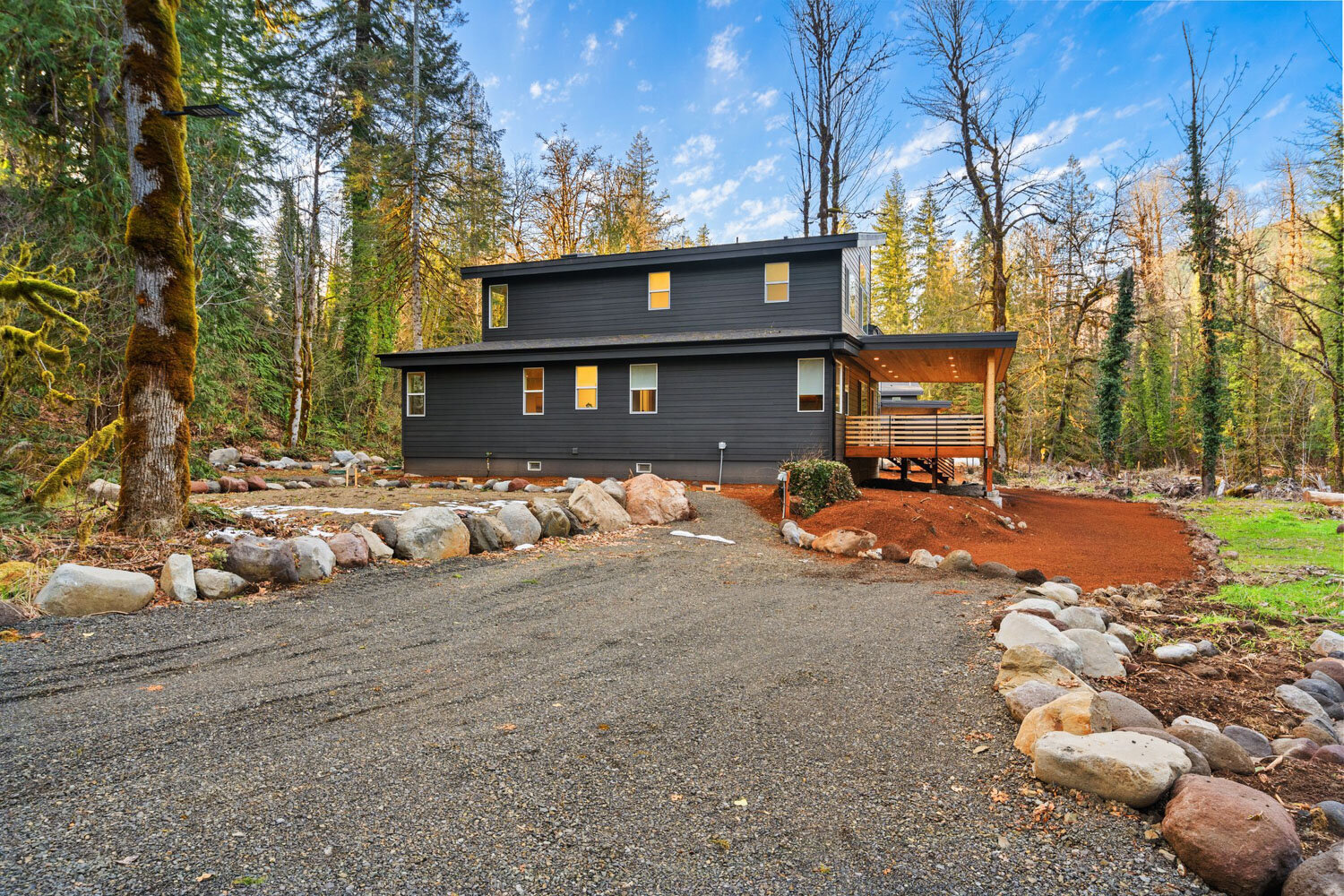 Modern black cabin surrounded by trees and gravel driveway.