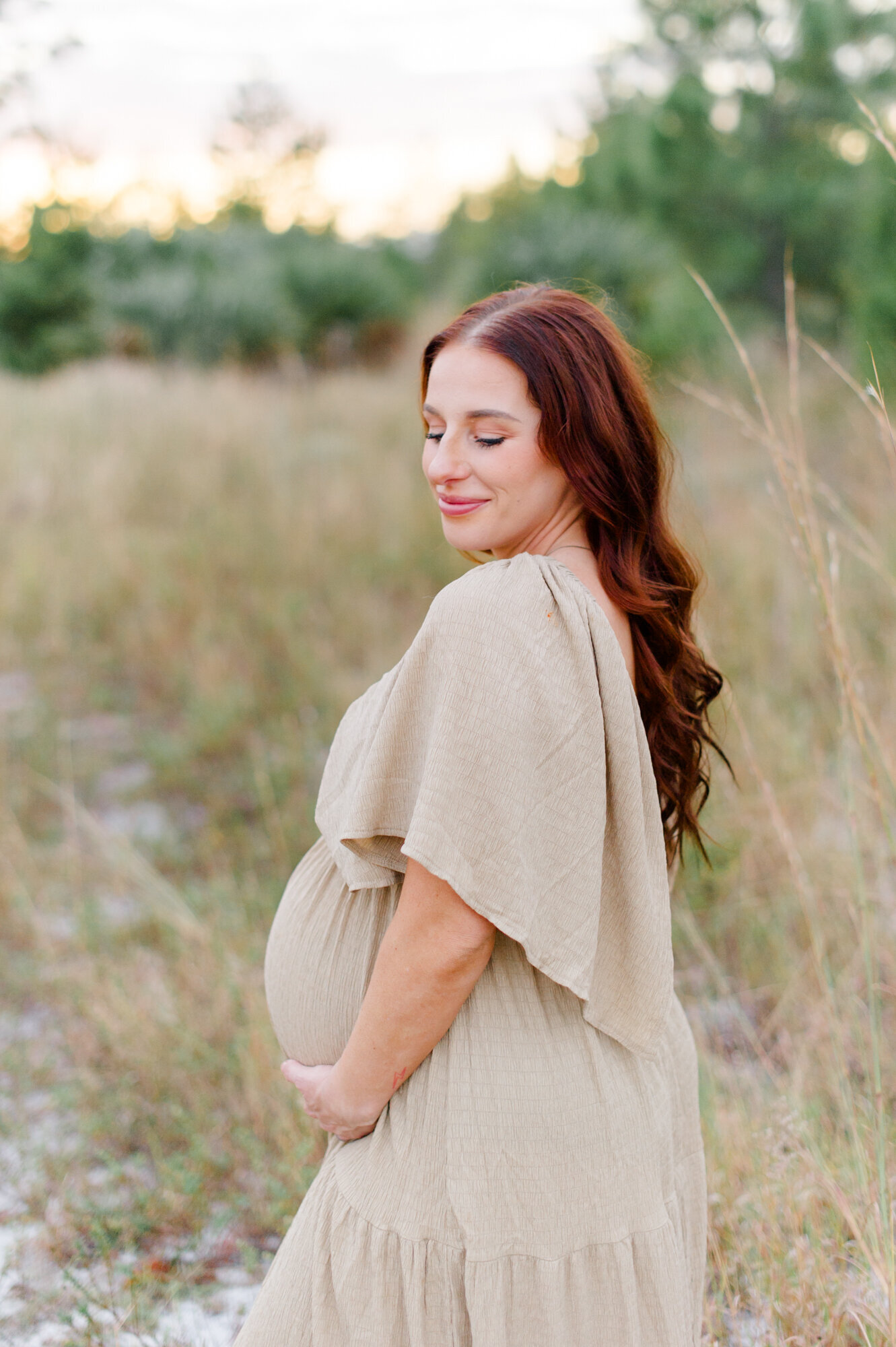 Pregnant mom stands in a tall grass field holding her belly during her sunset maternity session with an Orlando maternity photographer
